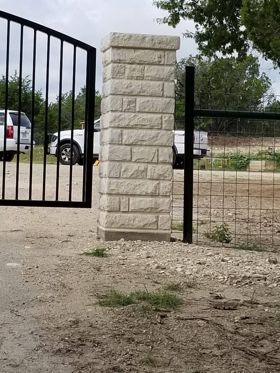 Closed black metal gate beside a stone pillar, with parked white vehicles behind it.