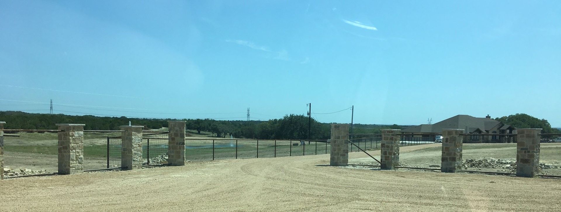 Wide view of a grassy field with stone gateposts, fence, and distant buildings under a clear blue sky