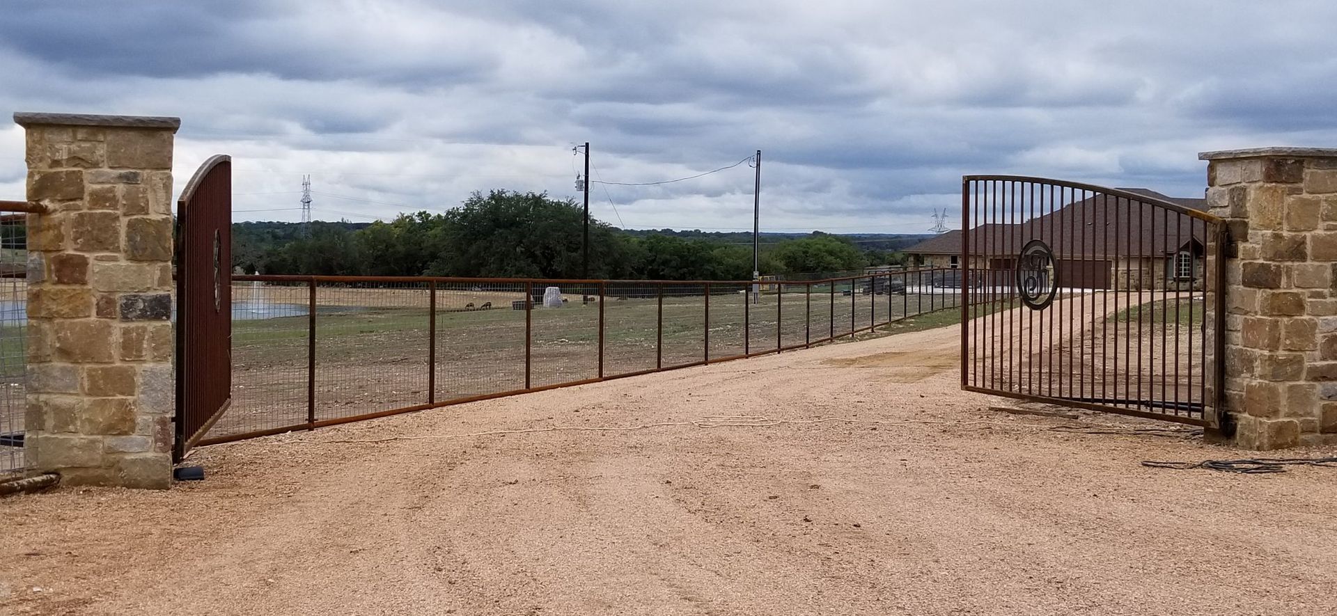 Open metal gate between stone pillars leading to a gravel driveway under cloudy skies