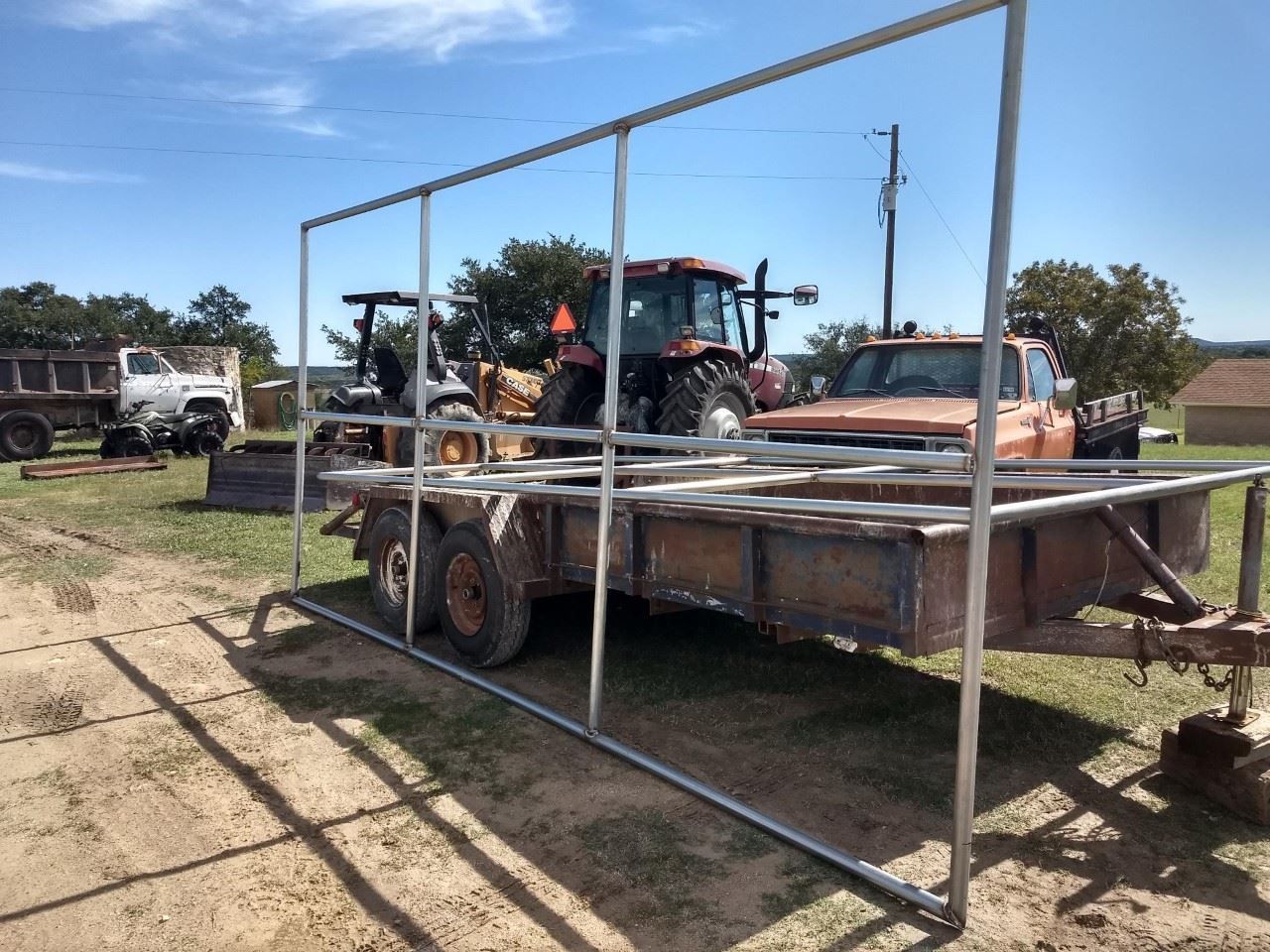 Flatbed trailer with metal frame in a dirt lot, carrying a tractor and equipment under a blue sky