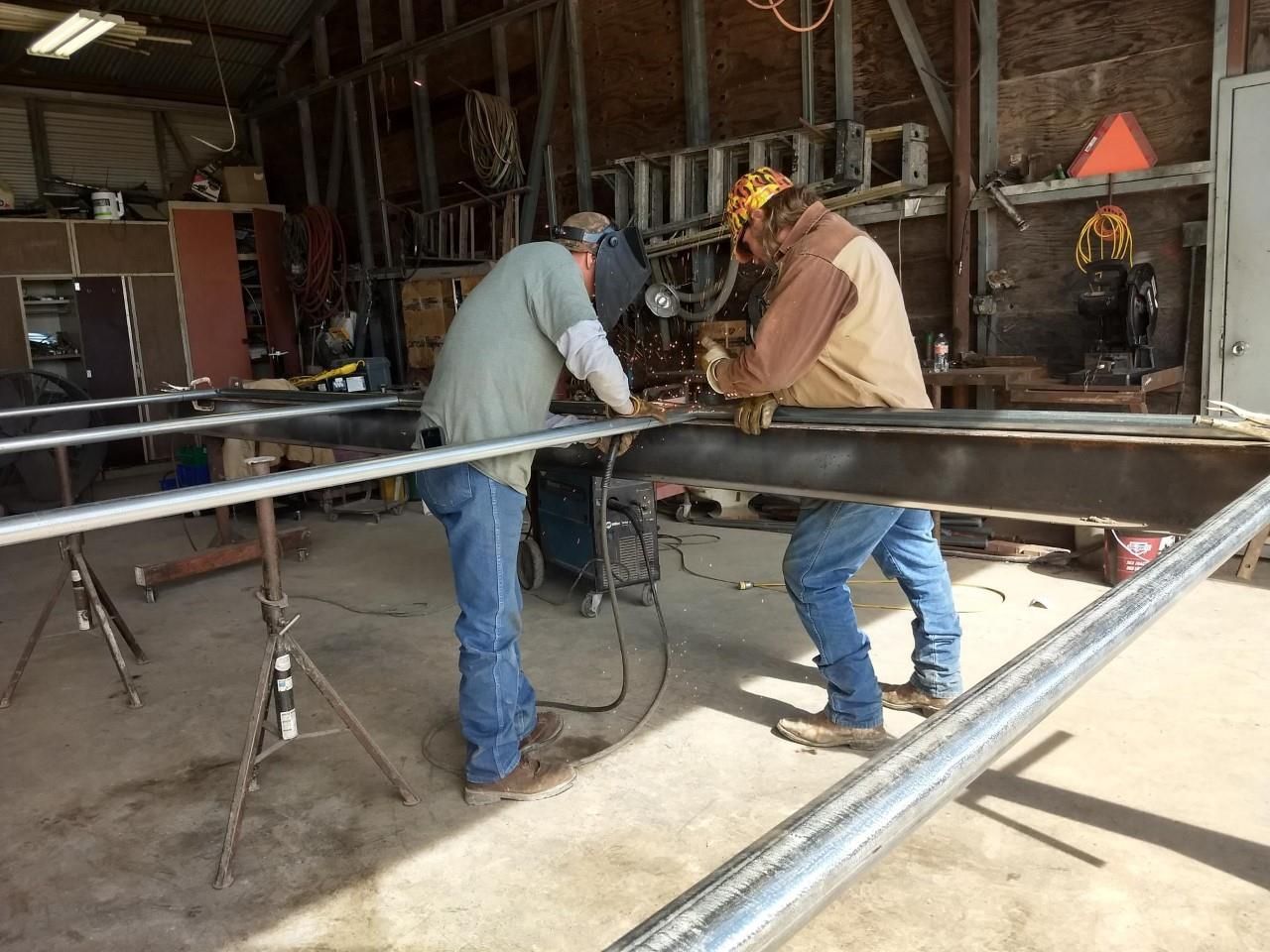 Two workers welding a metal frame in a workshop, sparks visible, with pipes and tools around them