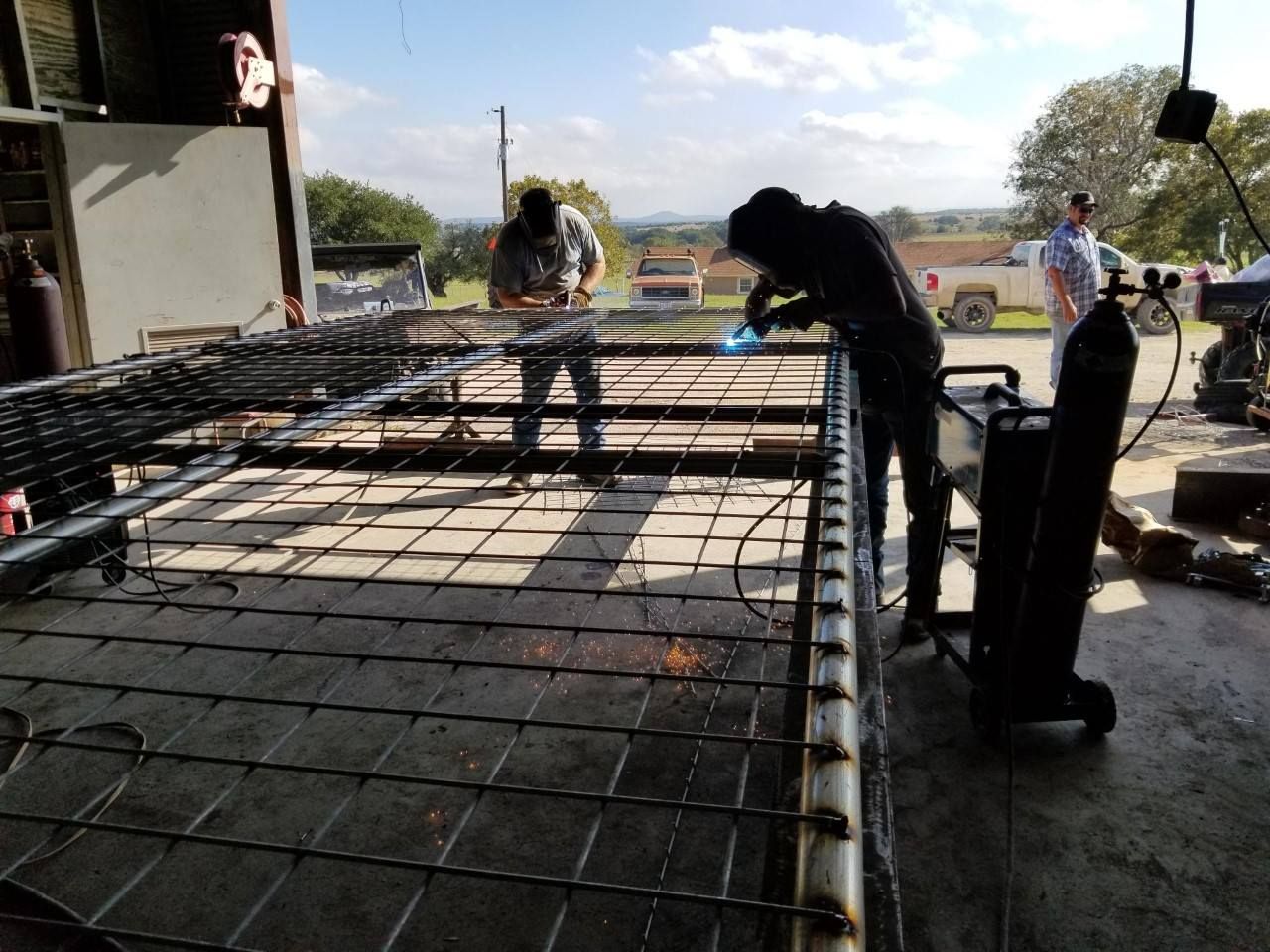 Workers weld a large metal rebar frame in an open-sided workshop