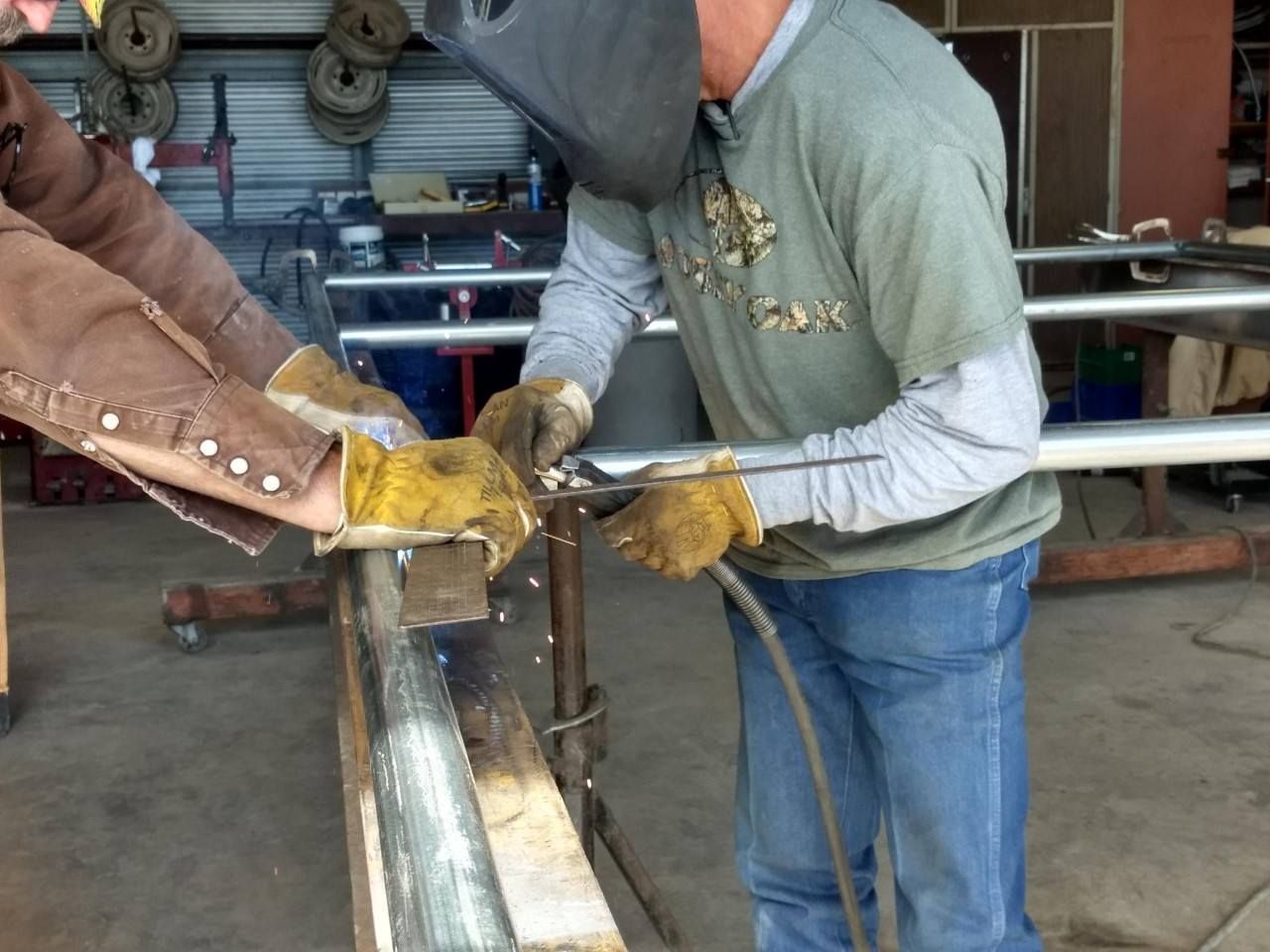 Two workers welding a metal frame in a workshop, wearing gloves and protective hoods.