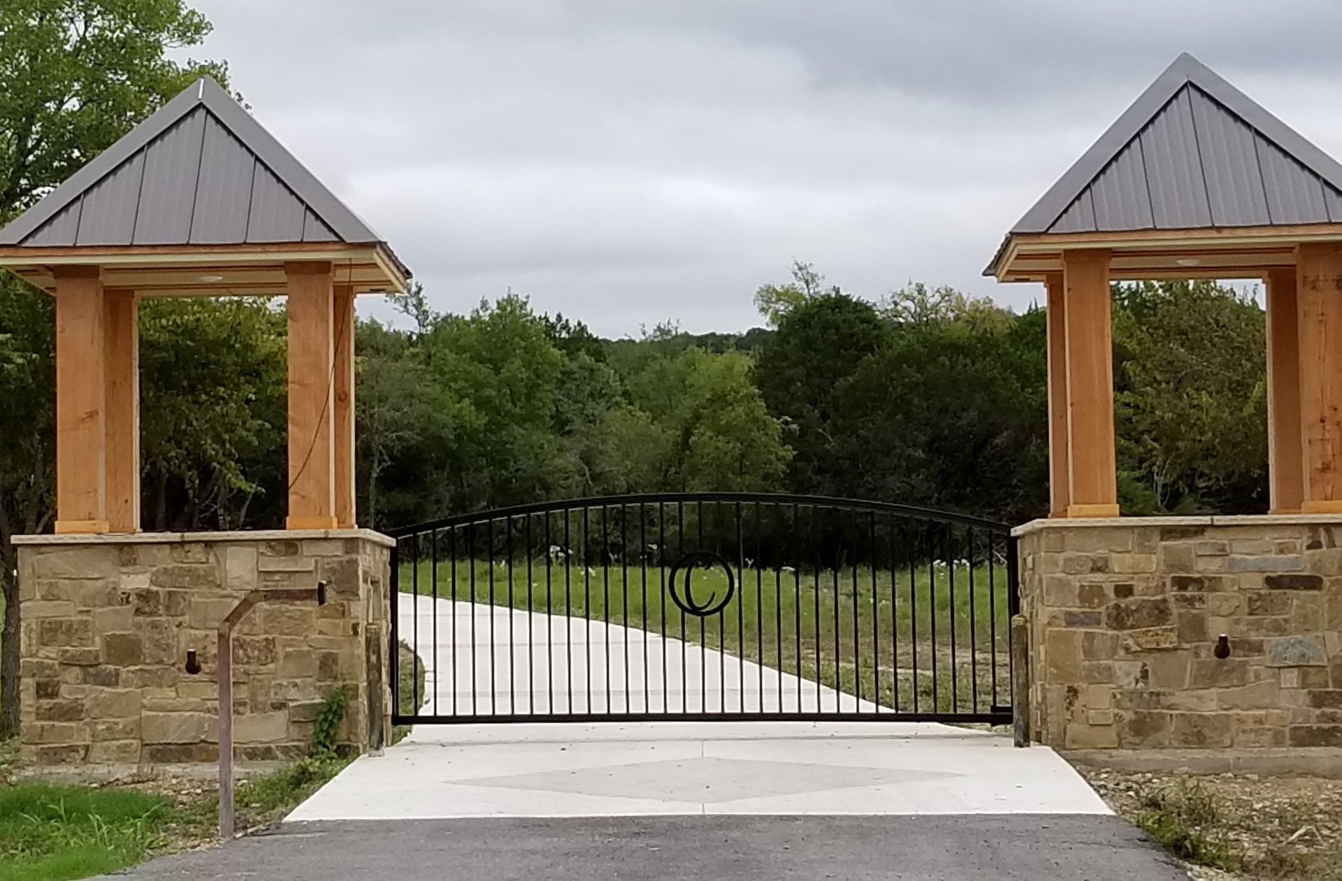 Stone gate entrance with black iron gate, flanked by stone pillars and wooden roofs, leading to a wooded drive