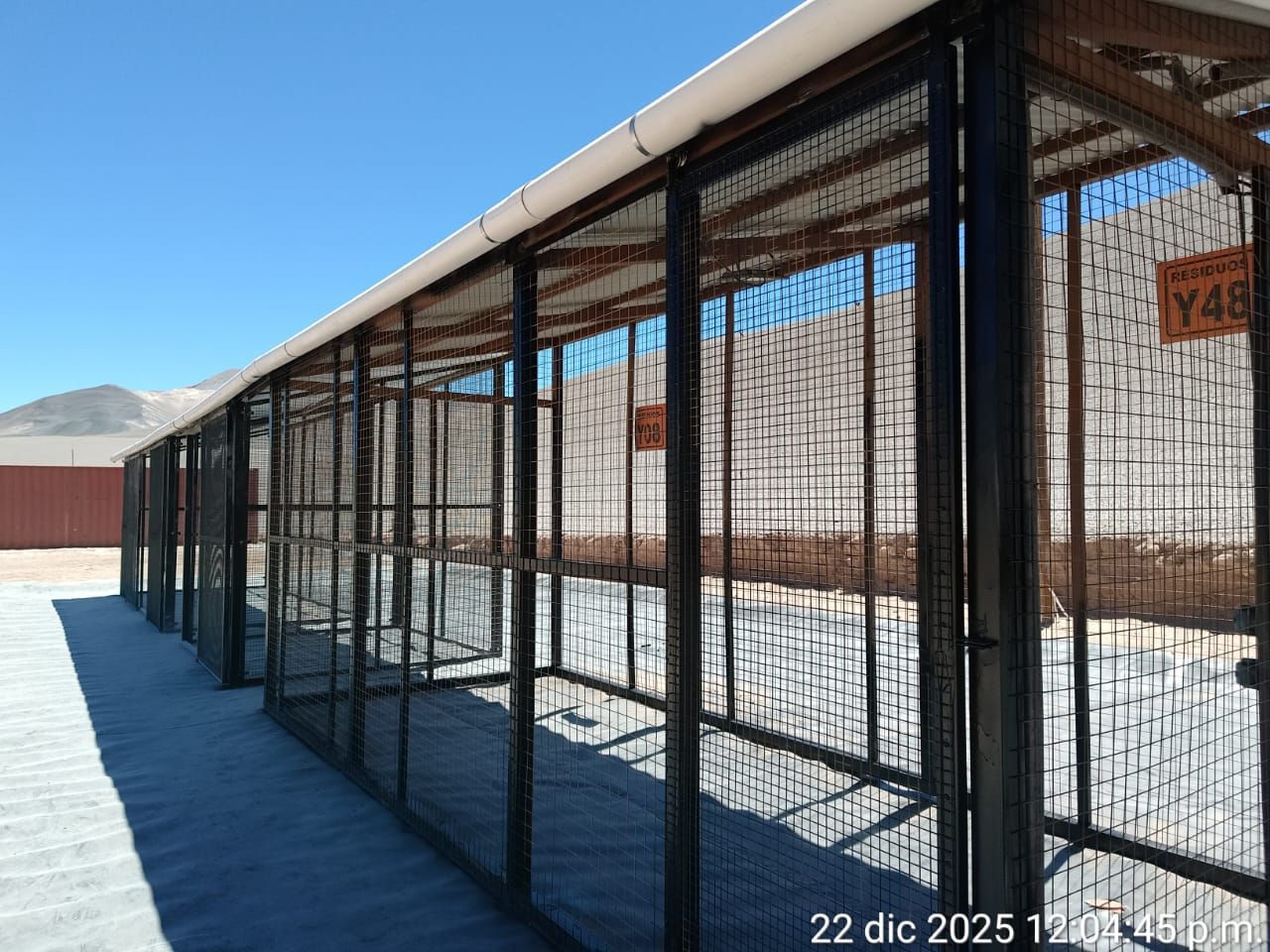 Largos corrales metálicos para animales con cercas de alambre en un patio nevado al aire libre, bajo un cielo azul claro.