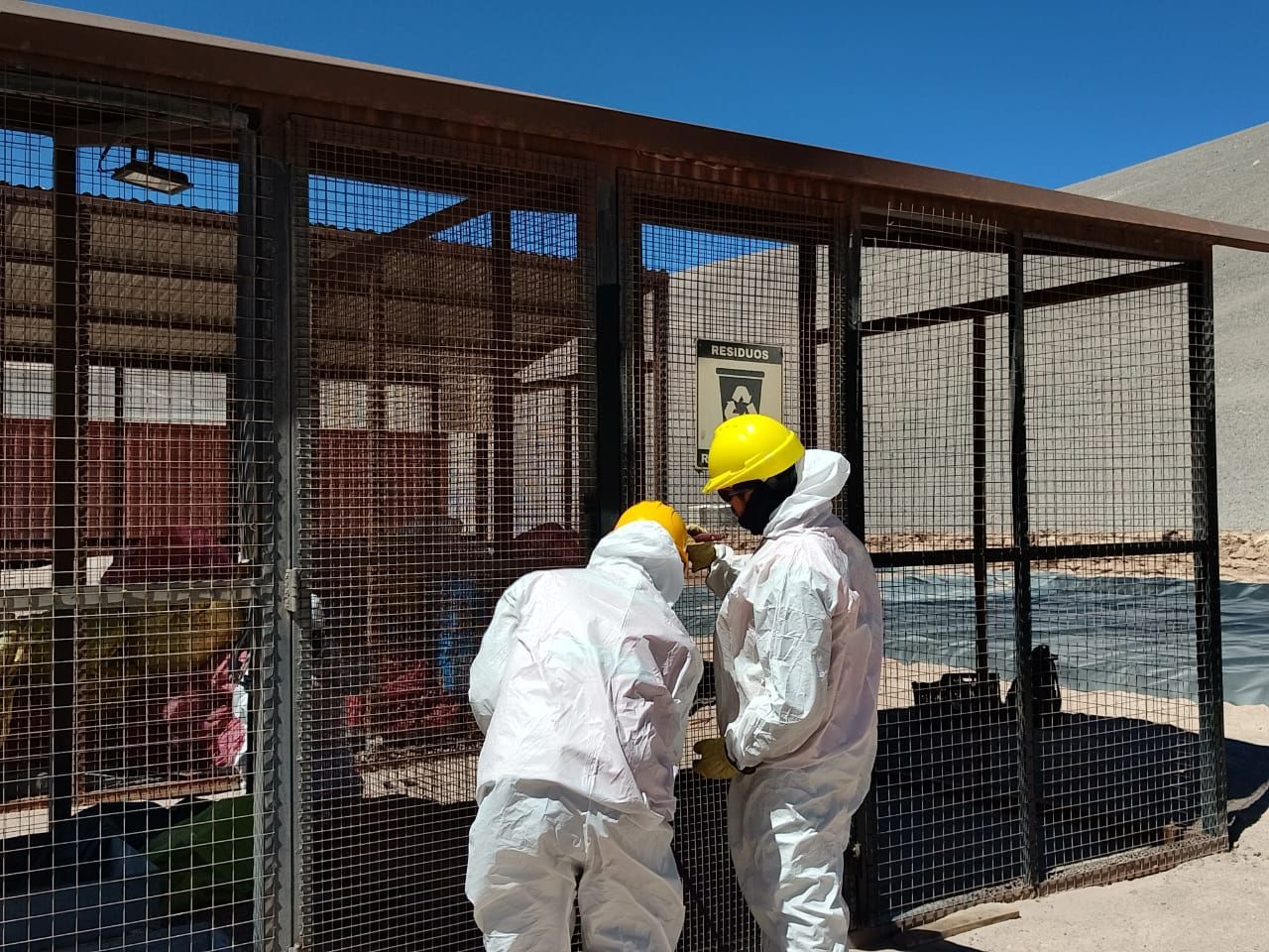 Dos trabajadores con trajes protectores blancos y cascos amarillos junto a una caja de cables en una obra de construcción.