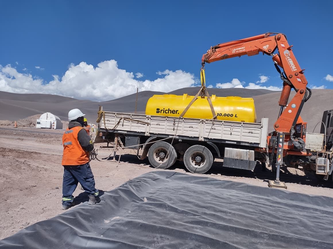 Obrero de la construcción cerca de una excavadora naranja cargando un camión cisterna amarillo en una obra de montaña nevada.