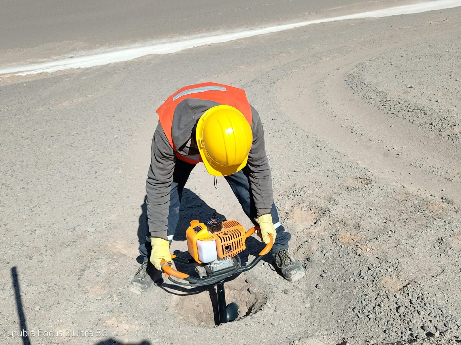 Obrero con casco amarillo utilizando un martillo neumático en la superficie de una carretera, visto desde arriba.