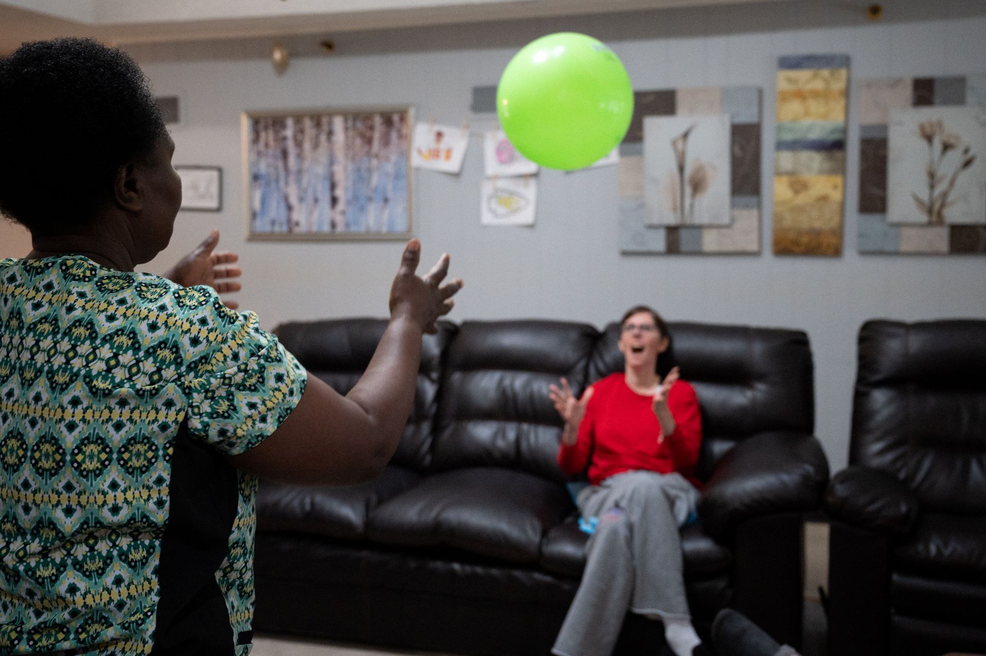 Two people tossing a bright green balloon in a living room with a black couch and wall art.
