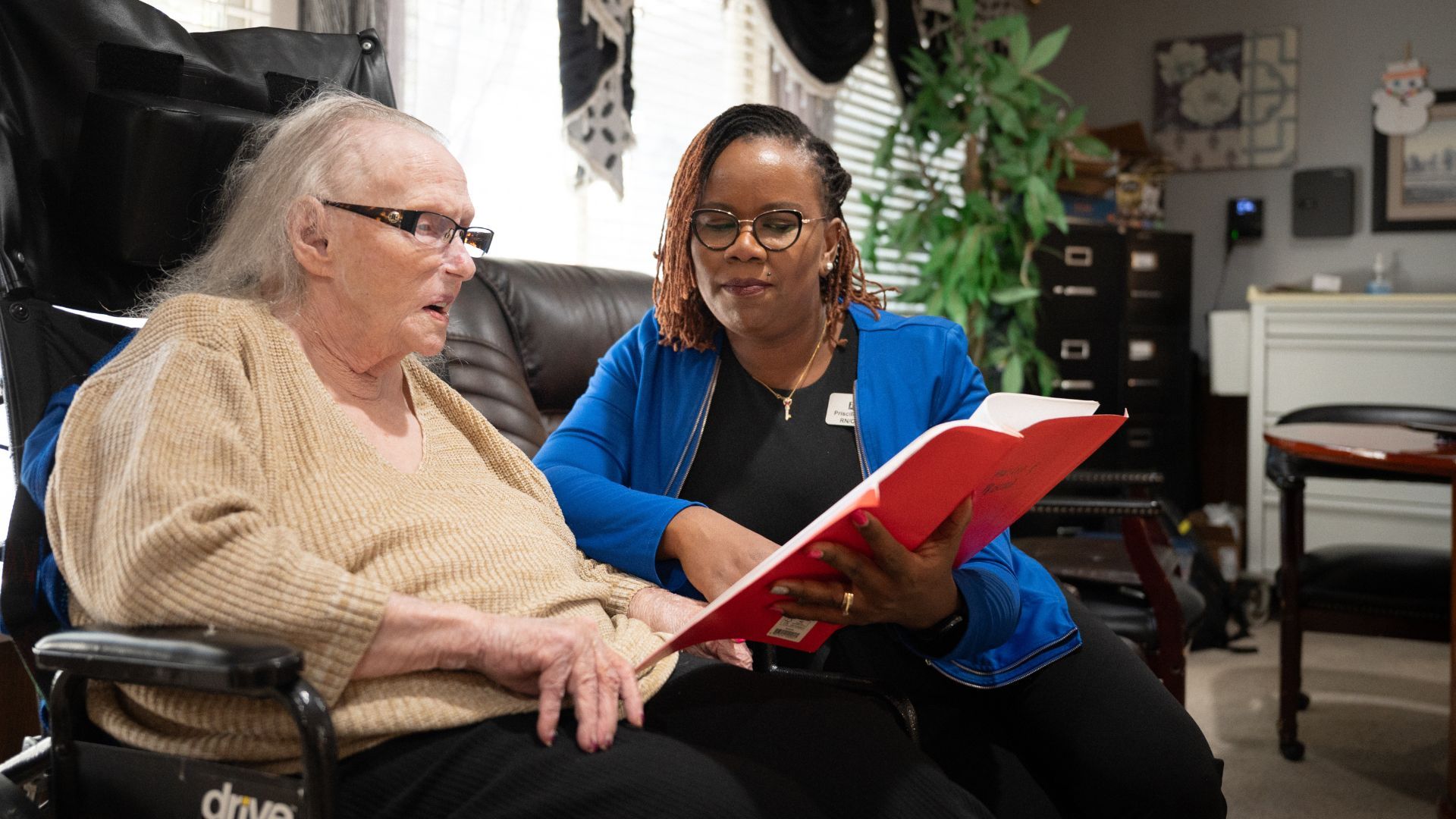 Two people sit indoors looking at a red folder, one in a wheelchair, in a cozy living room.