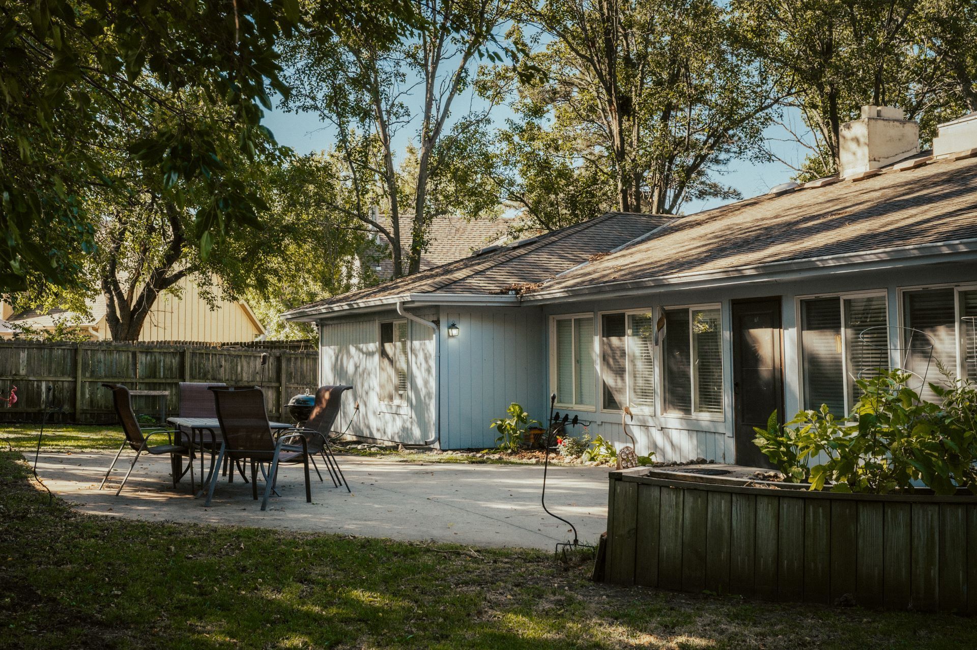 Backyard patio with table and chairs beside a light blue house under trees