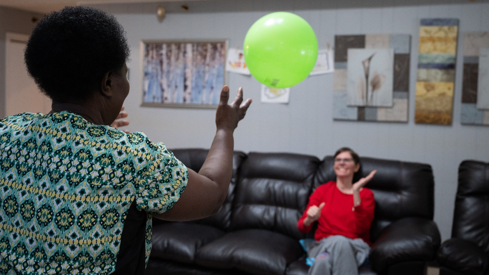 Person in patterned shirt tosses a green balloon toward someone seated on a black couch in a living room.