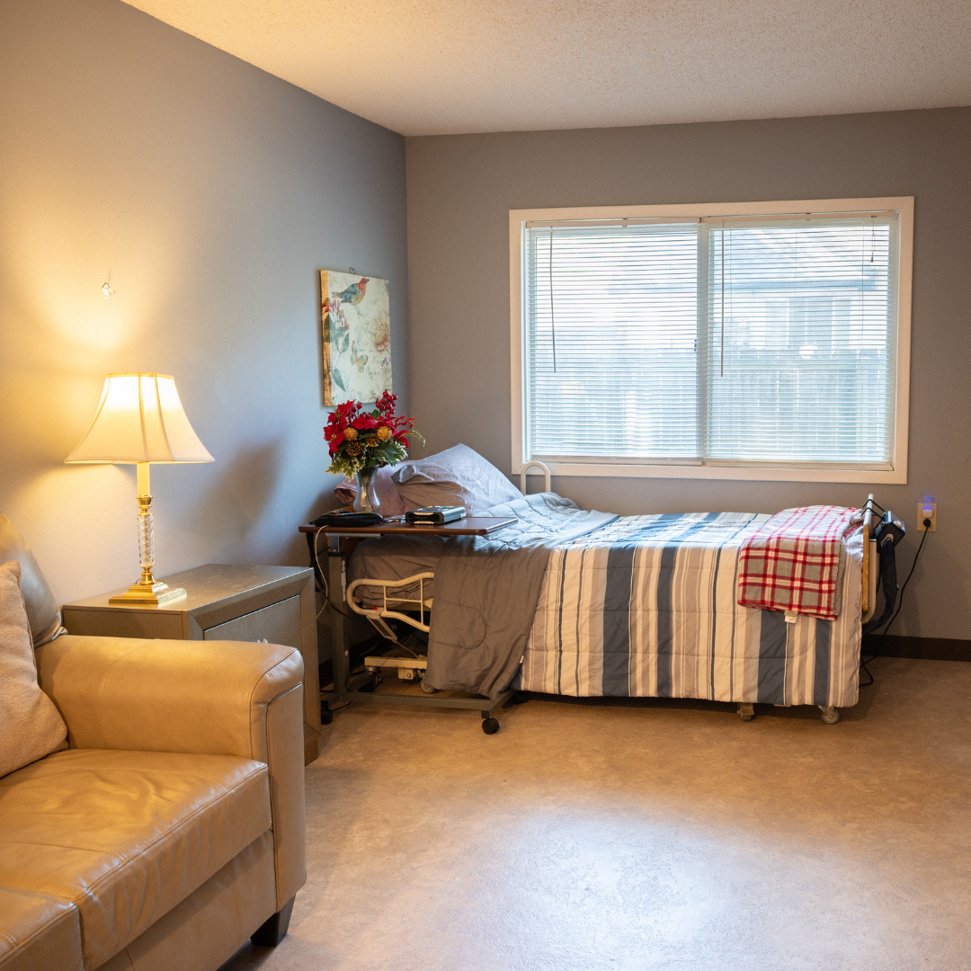 Cozy, sunlit room with a bed, couch, side table, and window. Gray walls, beige floor.