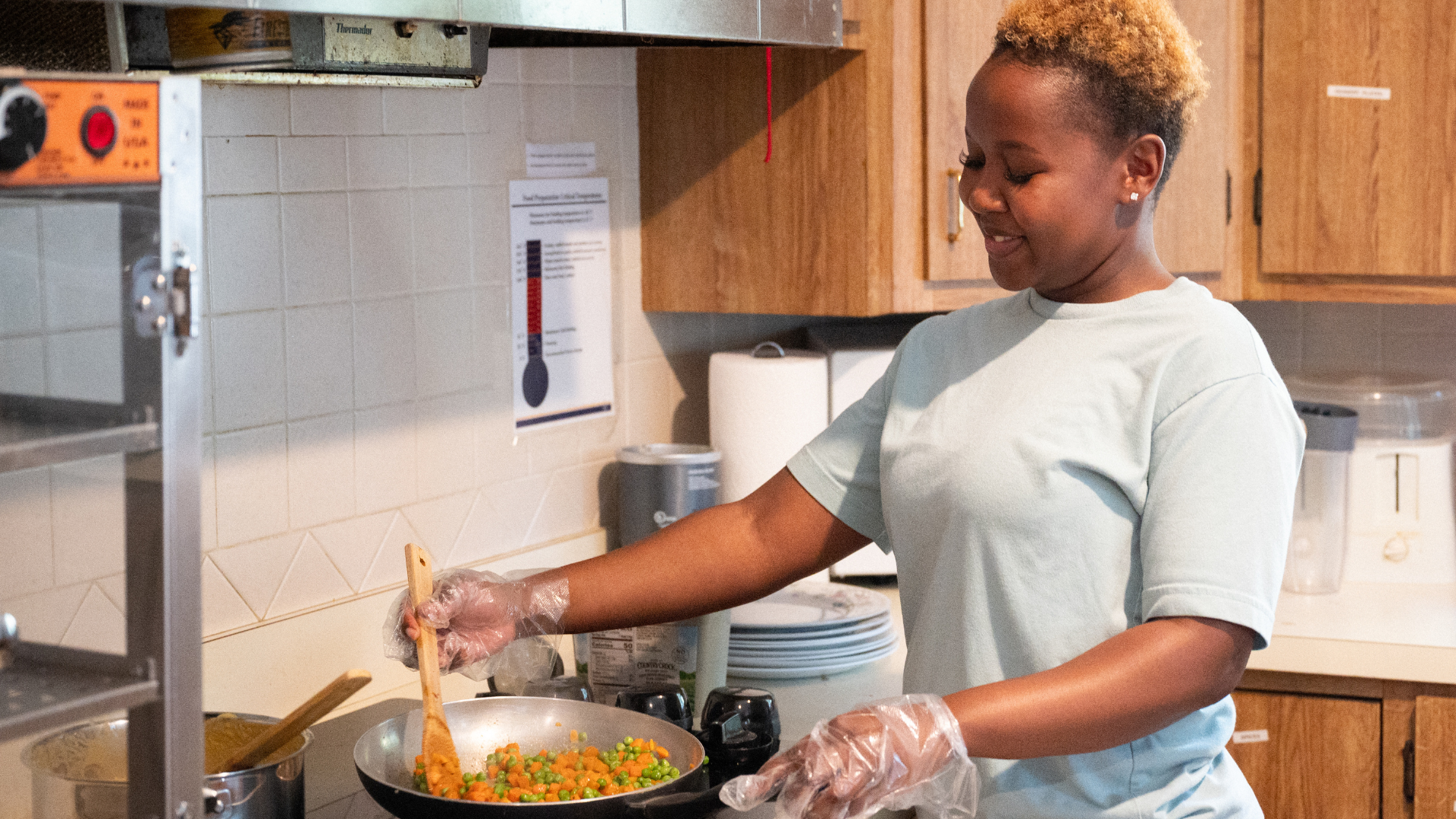 Person stirring food in a pan on a kitchen stove, wearing gloves and a light shirt.