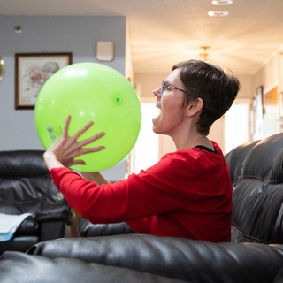 Woman in red shirt blows up green balloon indoors, smiling.