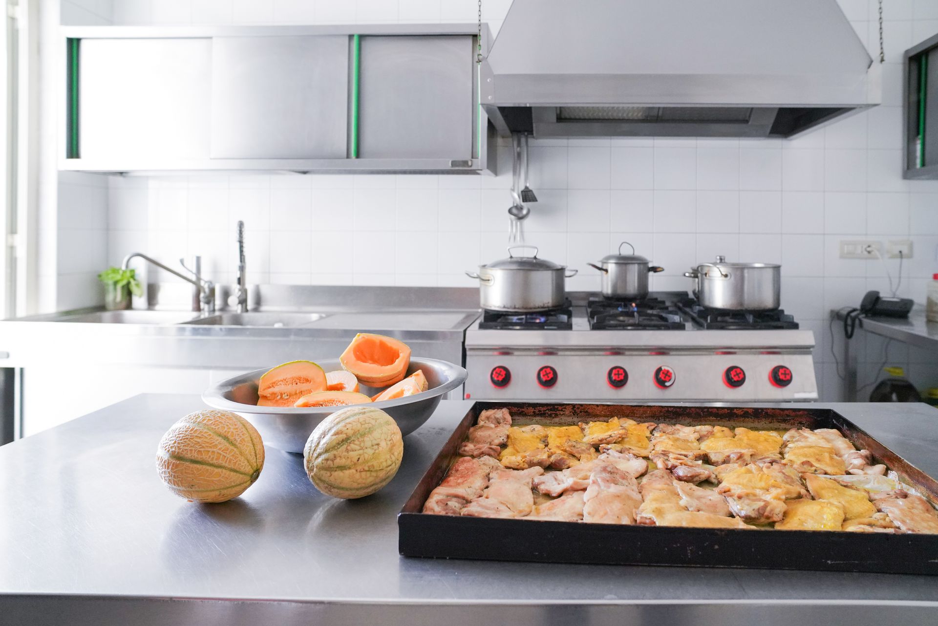 Cucina commerciale con superfici in acciaio inox, fornelli, teglie e meloni.