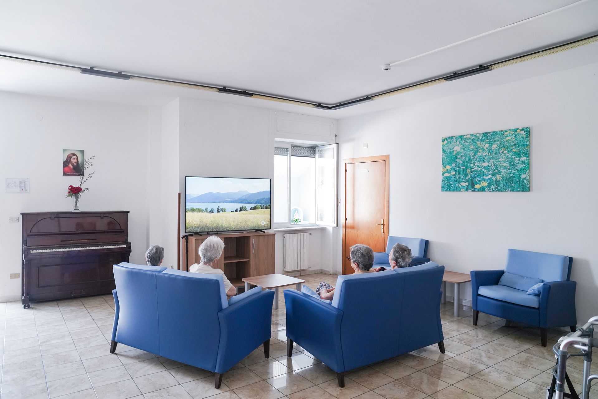 Anziani guardano la TV in un luminoso soggiorno con divani blu, un pianoforte e un quadro.