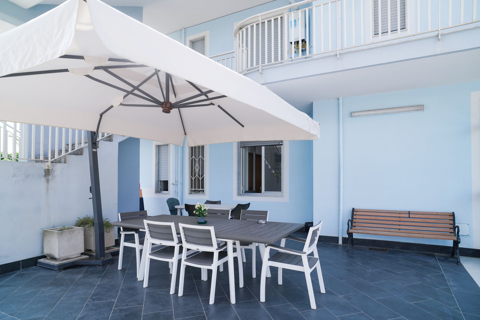 Patio con tavolo e sedie, ombreggiato da un ombrellone; edificio azzurro, panchina e balcone al secondo piano.