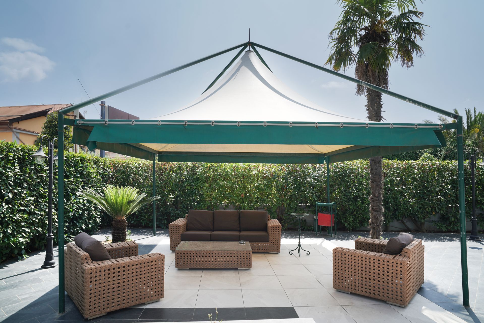 Area salotto all'aperto sotto una tettoia verde. Mobili in rattan sono disposti su un patio, con una siepe verde e una palma sullo sfondo.