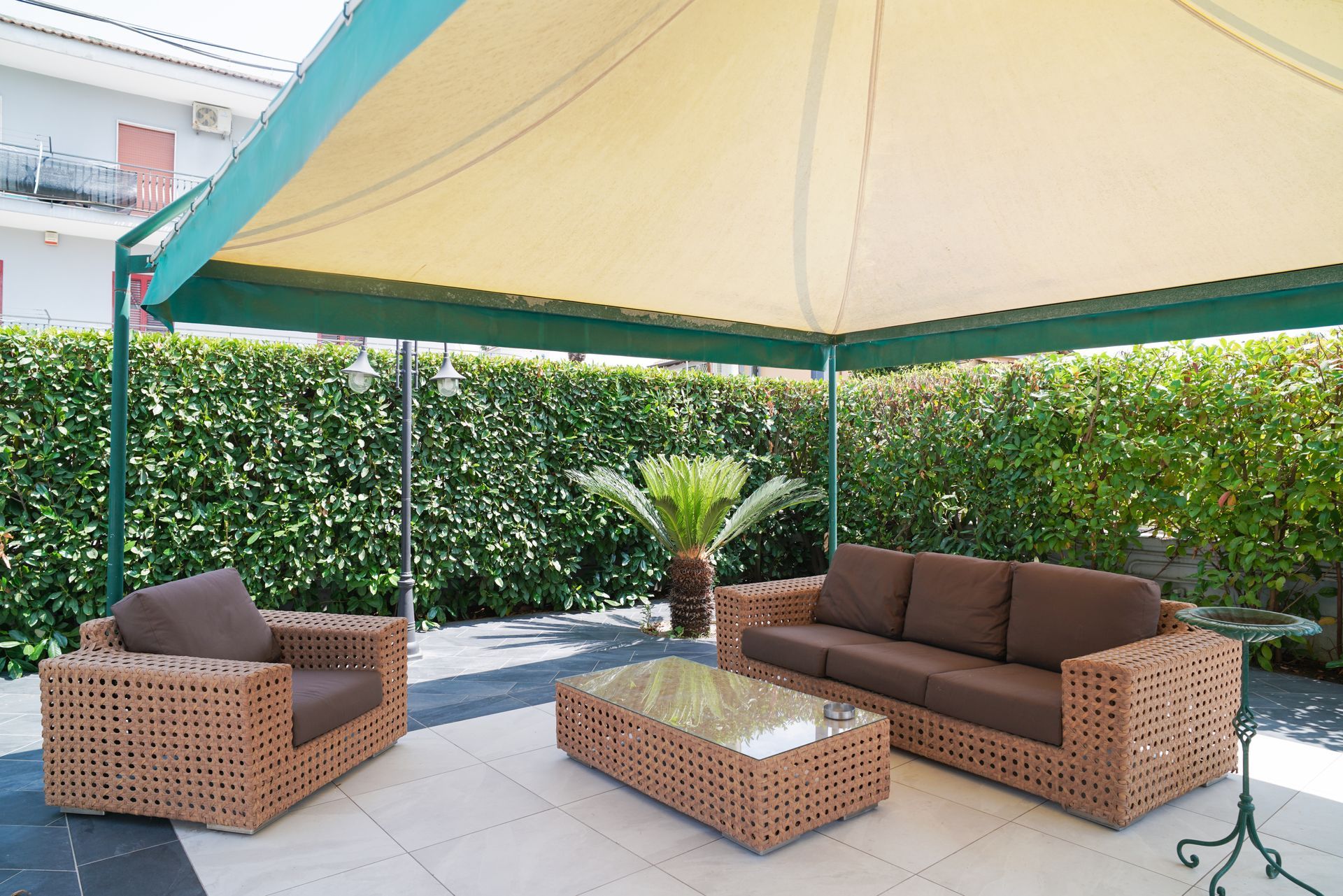 Patio con mobili in vimini sotto una tettoia. Siepe verde sullo sfondo.
