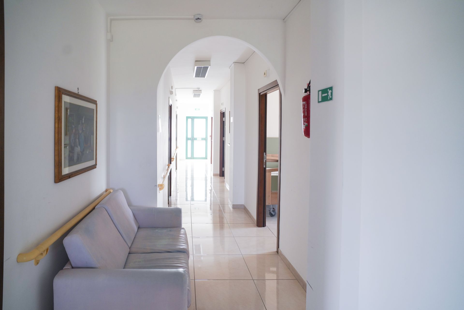 Lungo corridoio con pareti bianche, porta ad arco, divano e porte aperte.