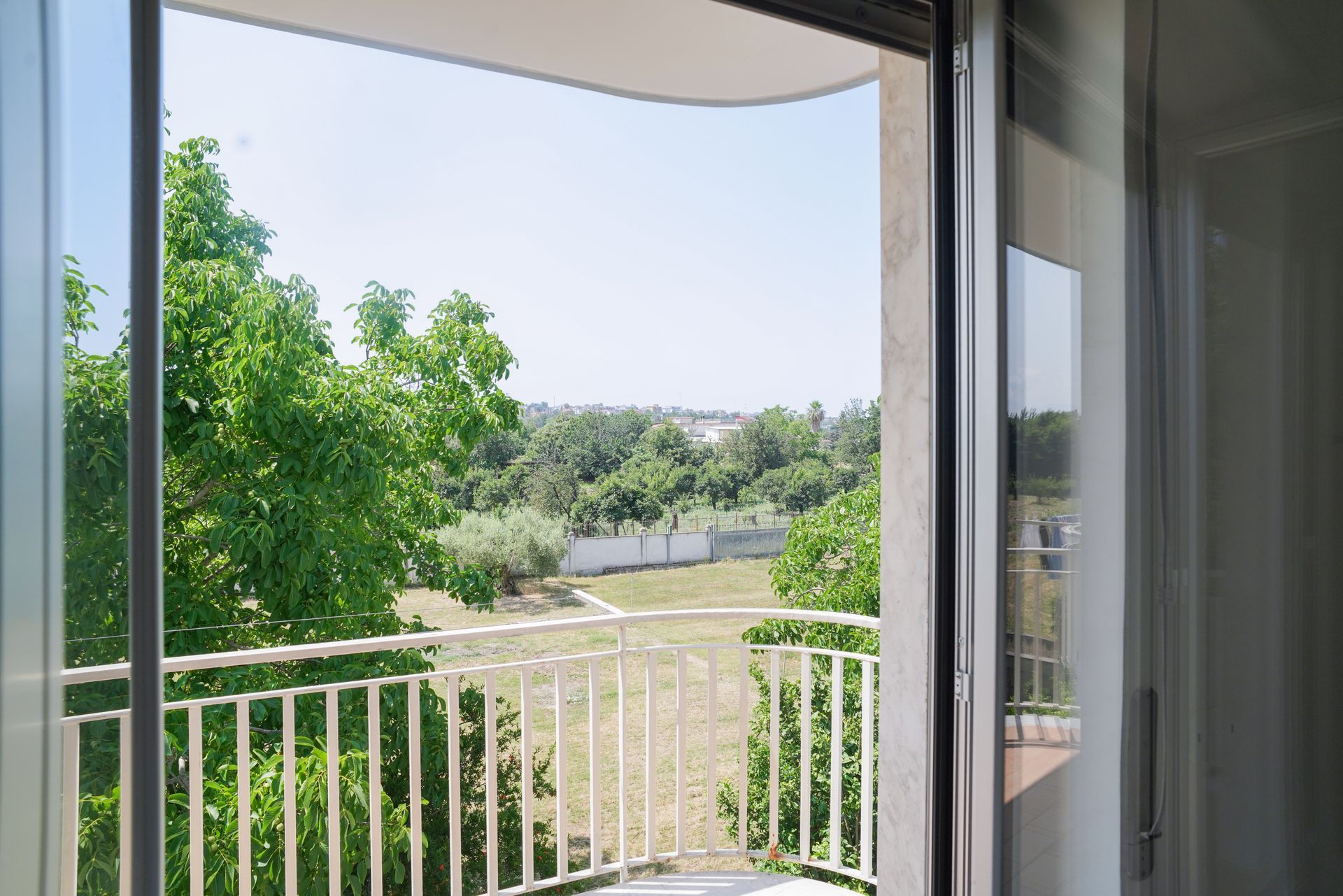 Vista da un balcone che si affaccia su un'area verde alberata, cielo azzurro.