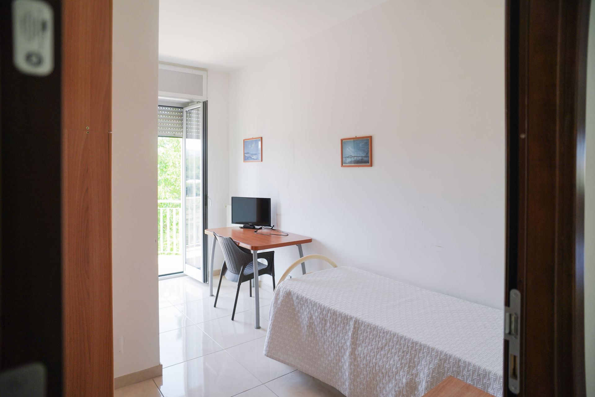 Vista su una semplice stanza con pareti bianche, letto, scrivania, sedia, piccola TV e porta-finestra.