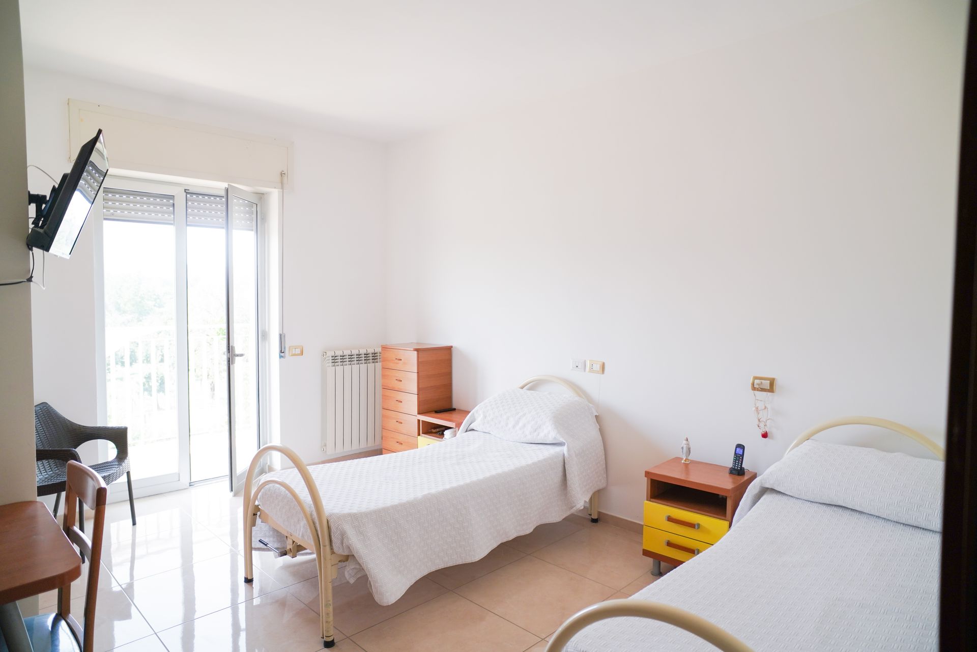 Due letti d'ospedale in una stanza luminosa con balcone e TV a parete.