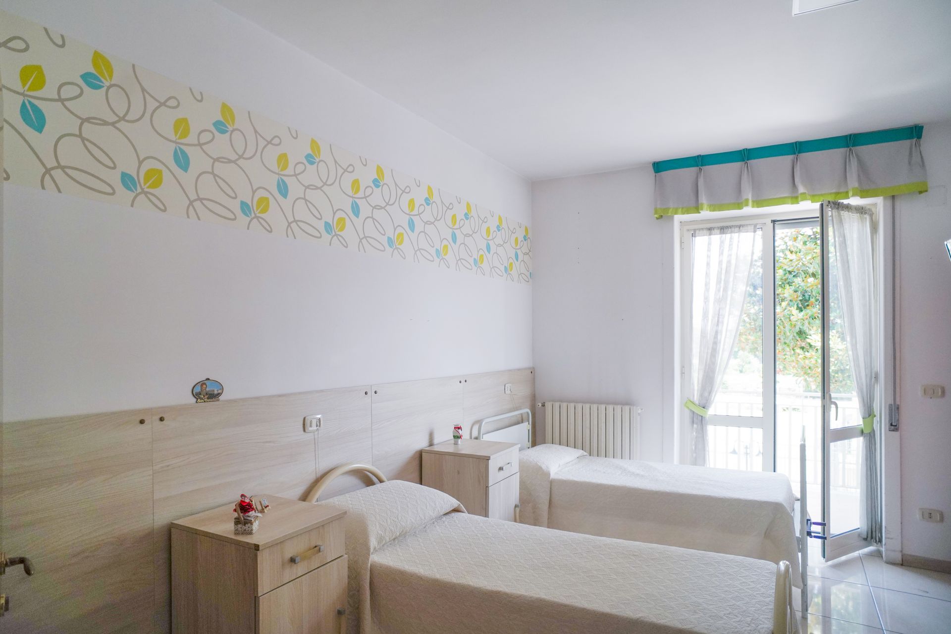 Due letti singoli in una stanza luminosa con pareti decorate con motivi floreali e porte-finestre.