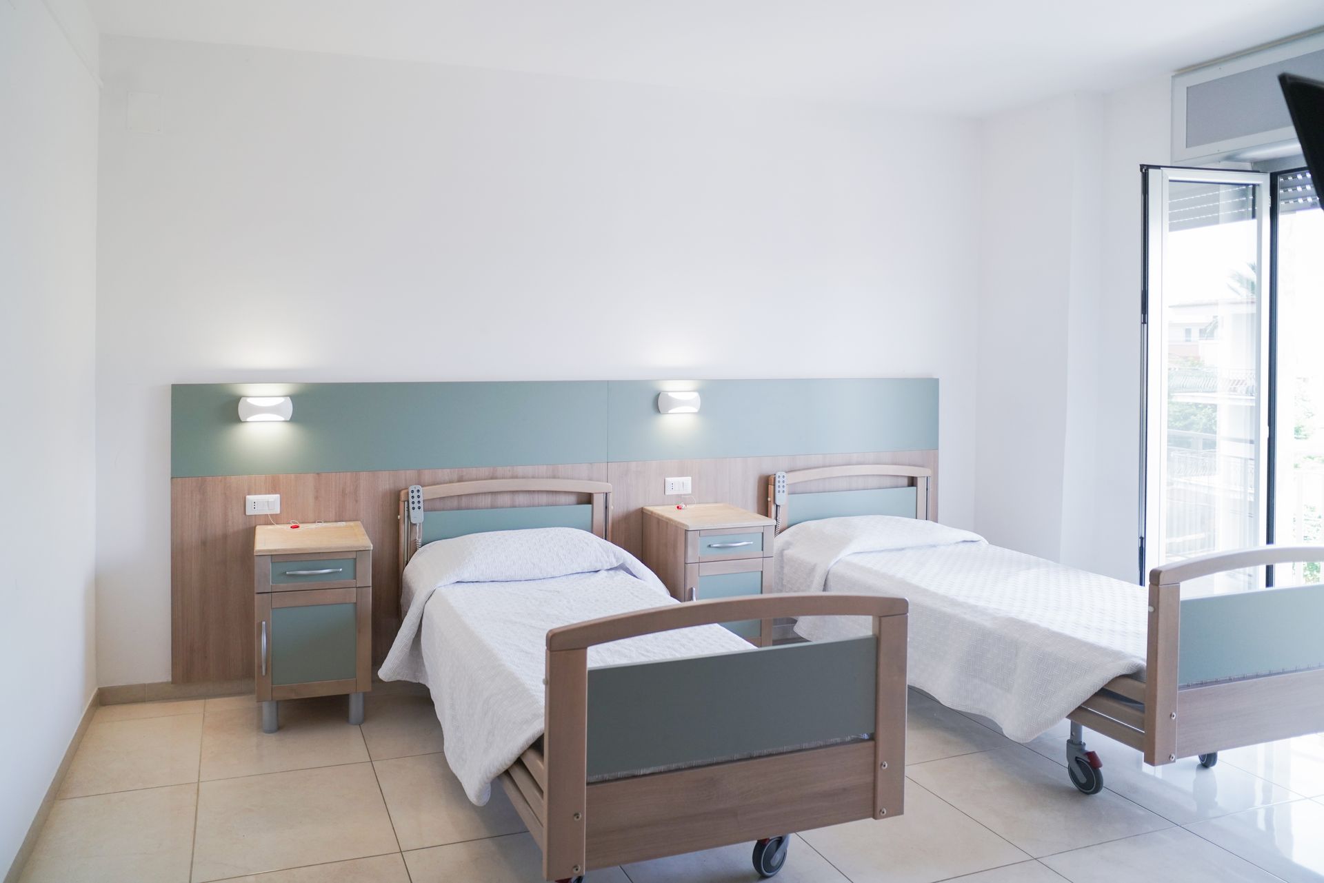Due letti d'ospedale in una stanza luminosa con comodini, una finestra e luci a parete.