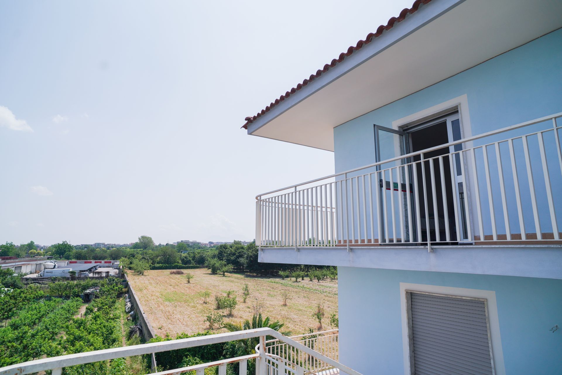 Casa blu con balcone che si affaccia su un paesaggio rurale con campi aperti e alberi verdi.
