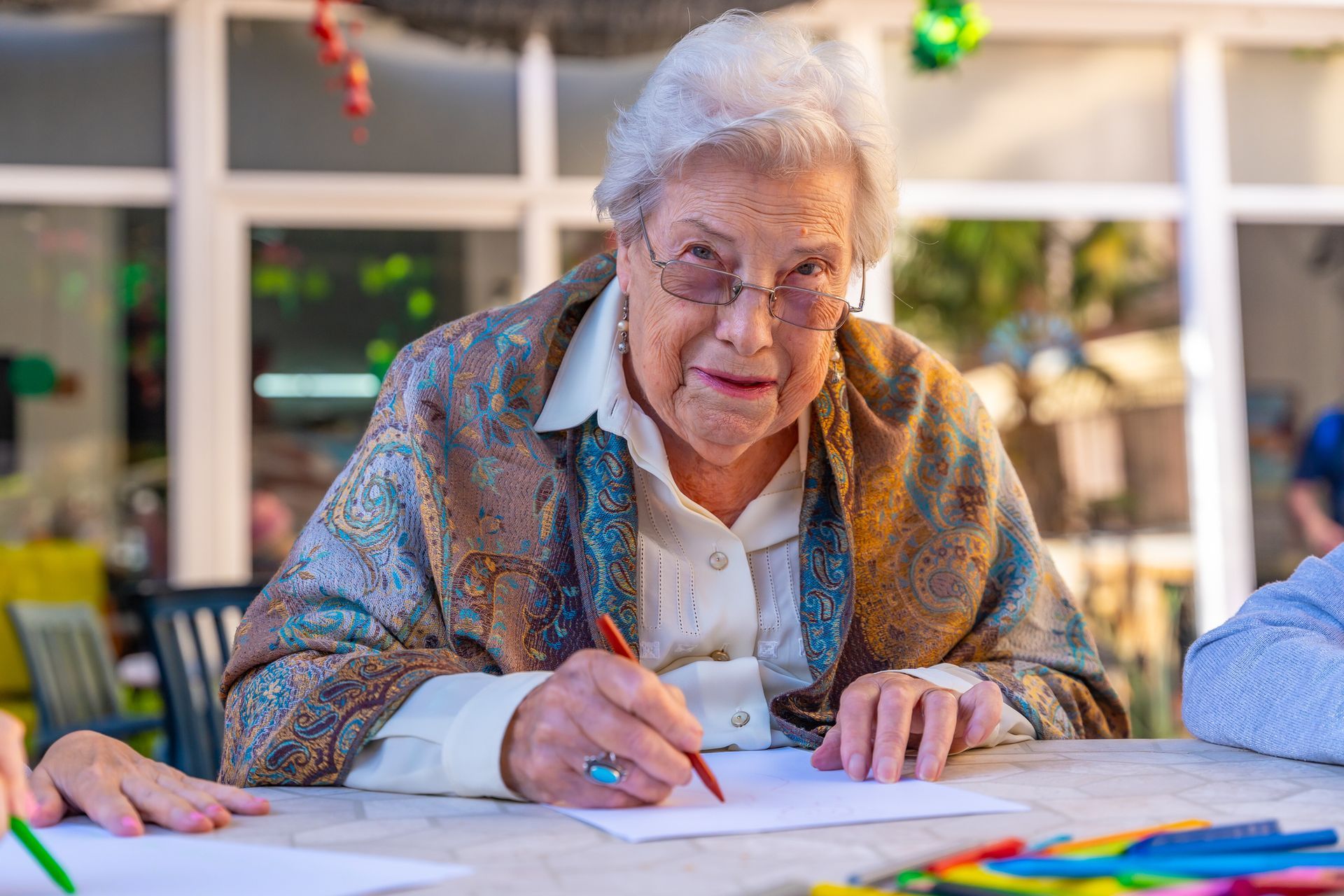 Donna anziana con occhiali e scialle, che disegna con una penna rossa all'aperto.