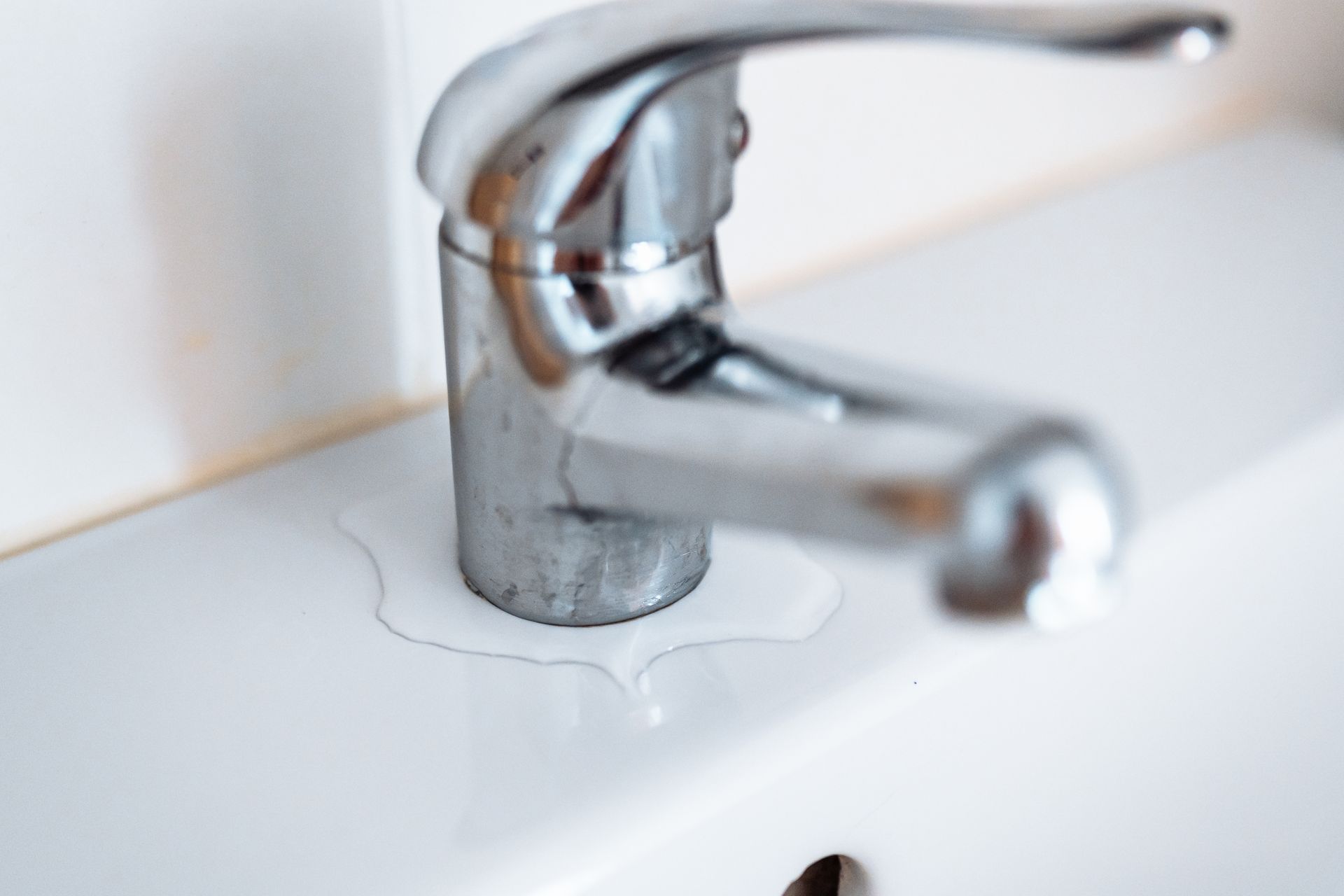 A close-up of a silver bathroom faucet with a pool of water leaking onto the white ceramic sink base.