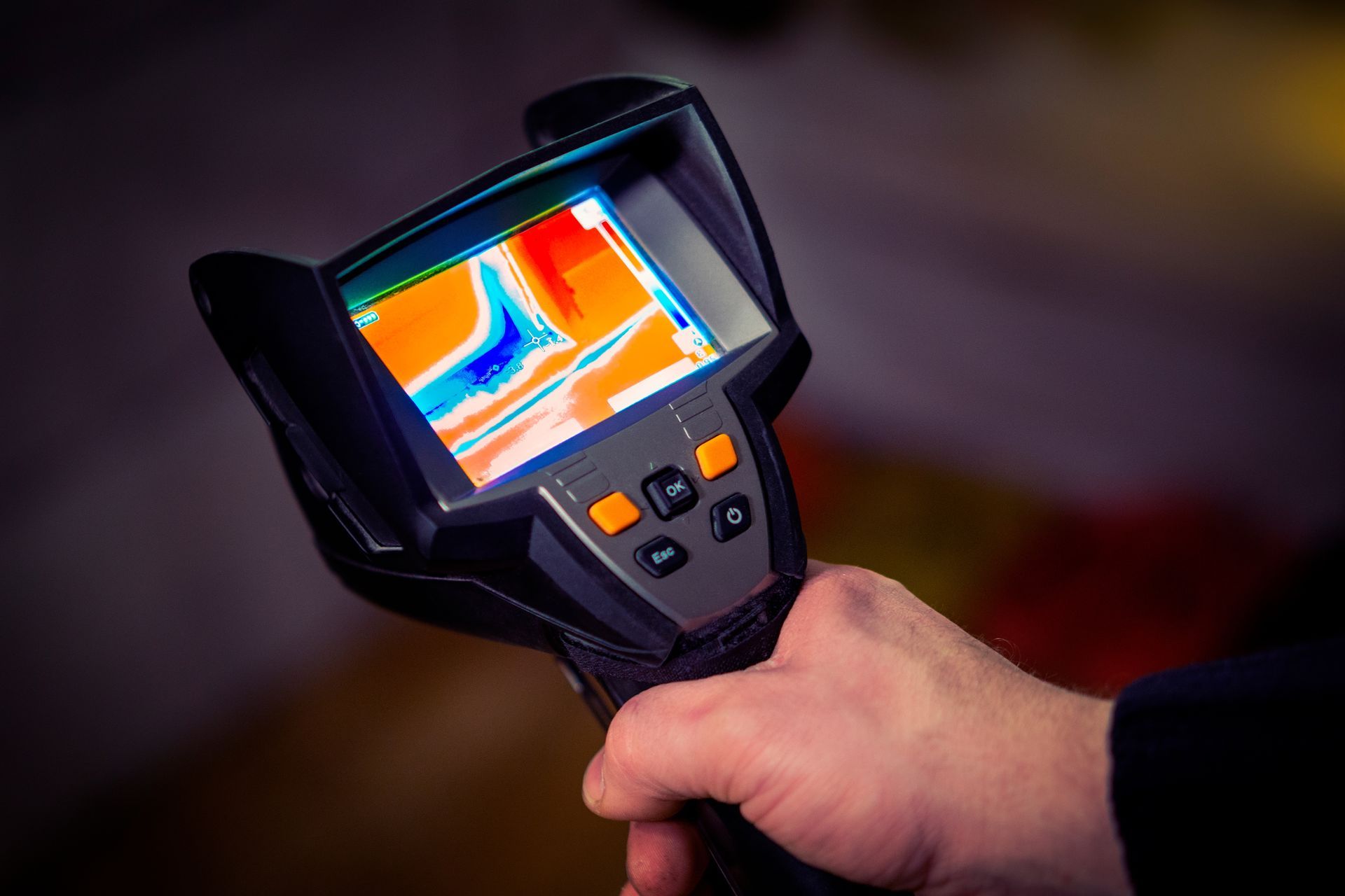 A hand holds a thermal imaging camera displaying a color-coded heat map of a surface.