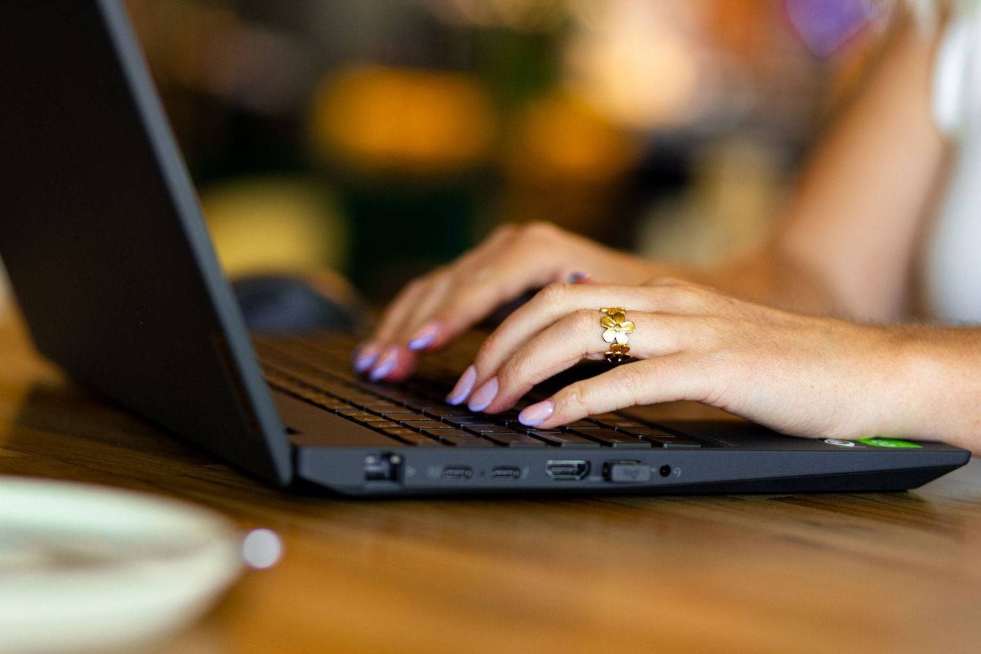 Handen typen op een zwarte laptop aan een houten tafel, met een wazige, warme achtergrond.