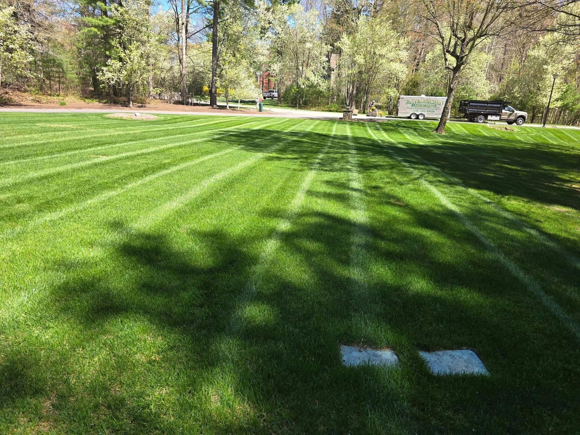 A lush, green lawn with distinct mowing stripes leading toward trees, a parked trailer, and a truck in the distance.