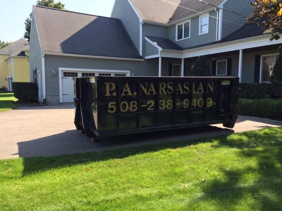 A black dumpster with company name and contact number parked on a suburban house driveway.