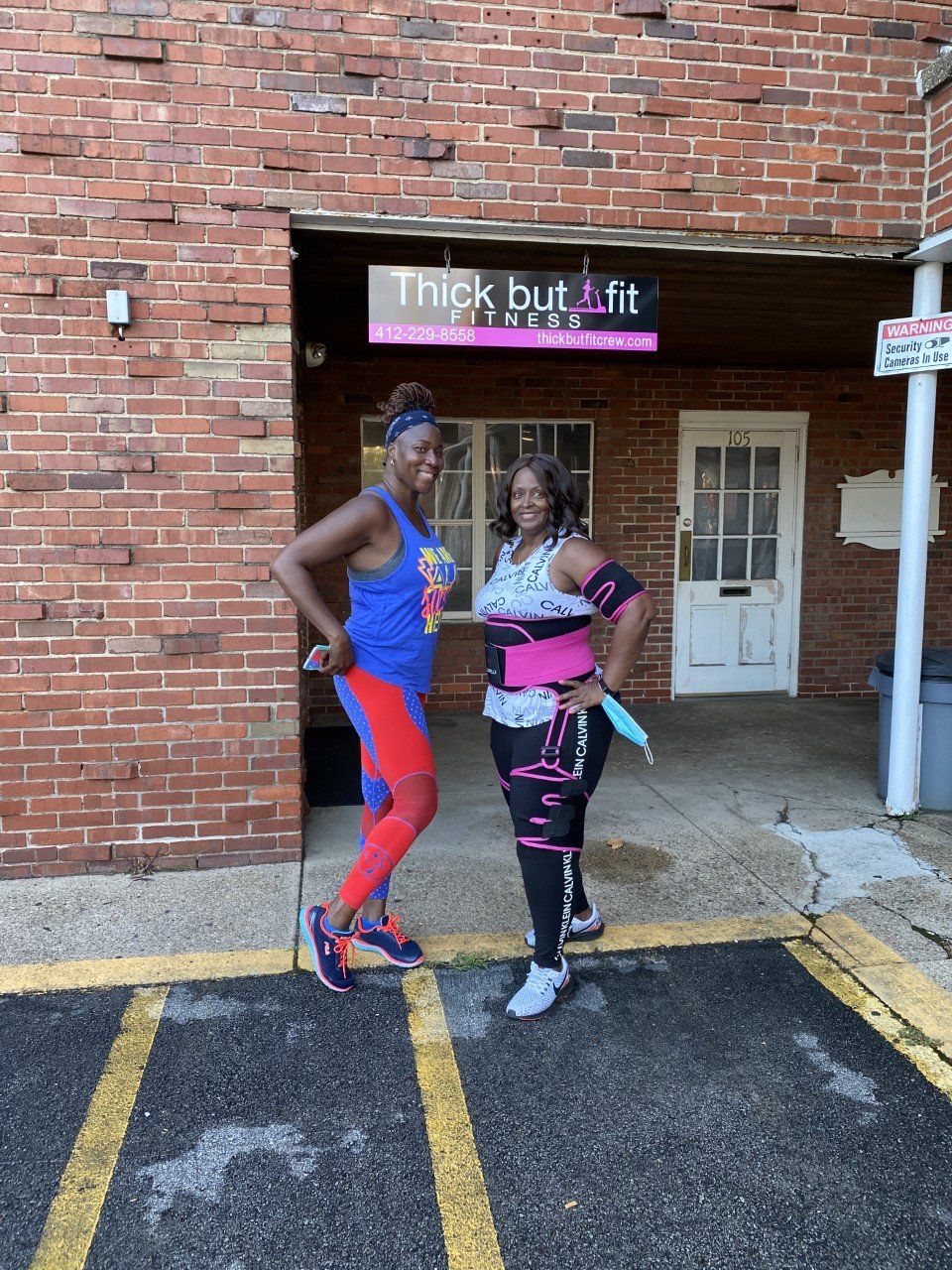 Two people posing outside a brick building with a “Thick but fit” sign above the door.