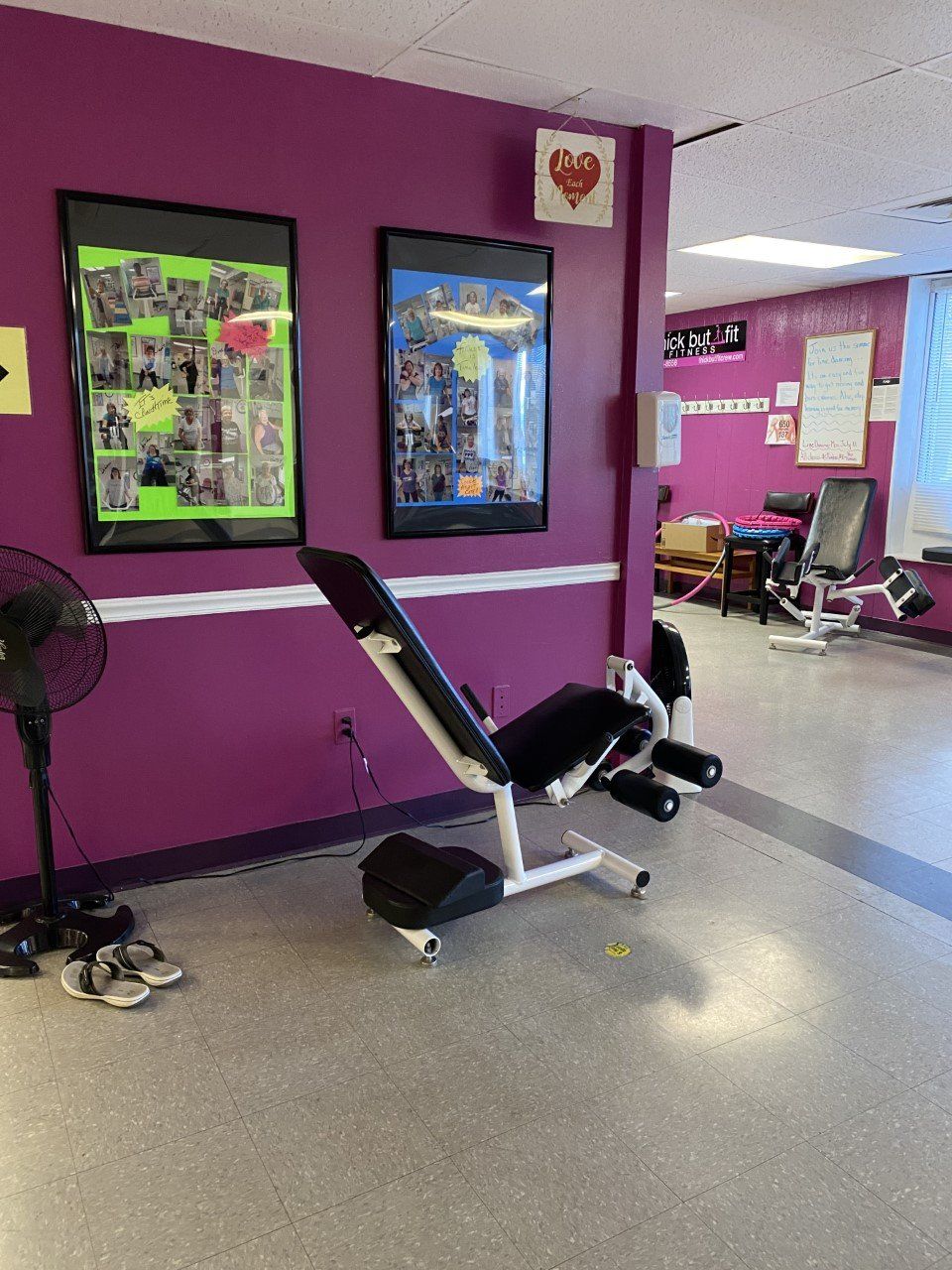 Gym with purple wall, bench press machine, framed posters, and a yoga mat in a fitness area.