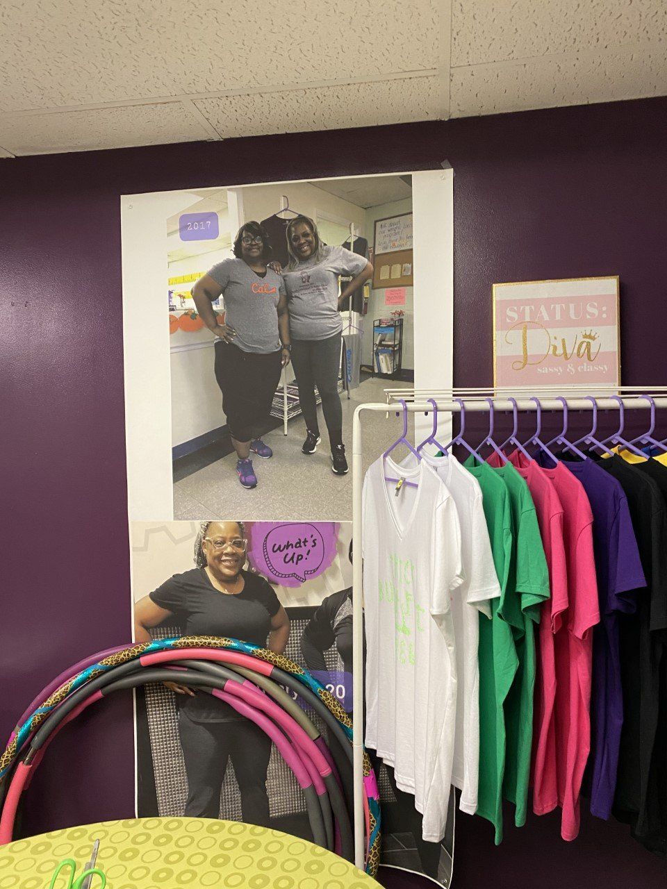 Clothing store display with colorful shirts on a rack and a poster of people shopping on a purple wall.