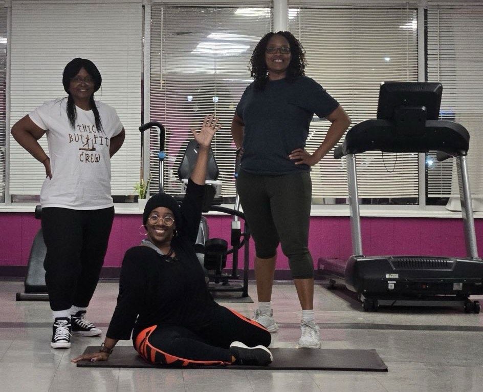 Three people posing in a gym, with one seated on a mat and two standing behind her.