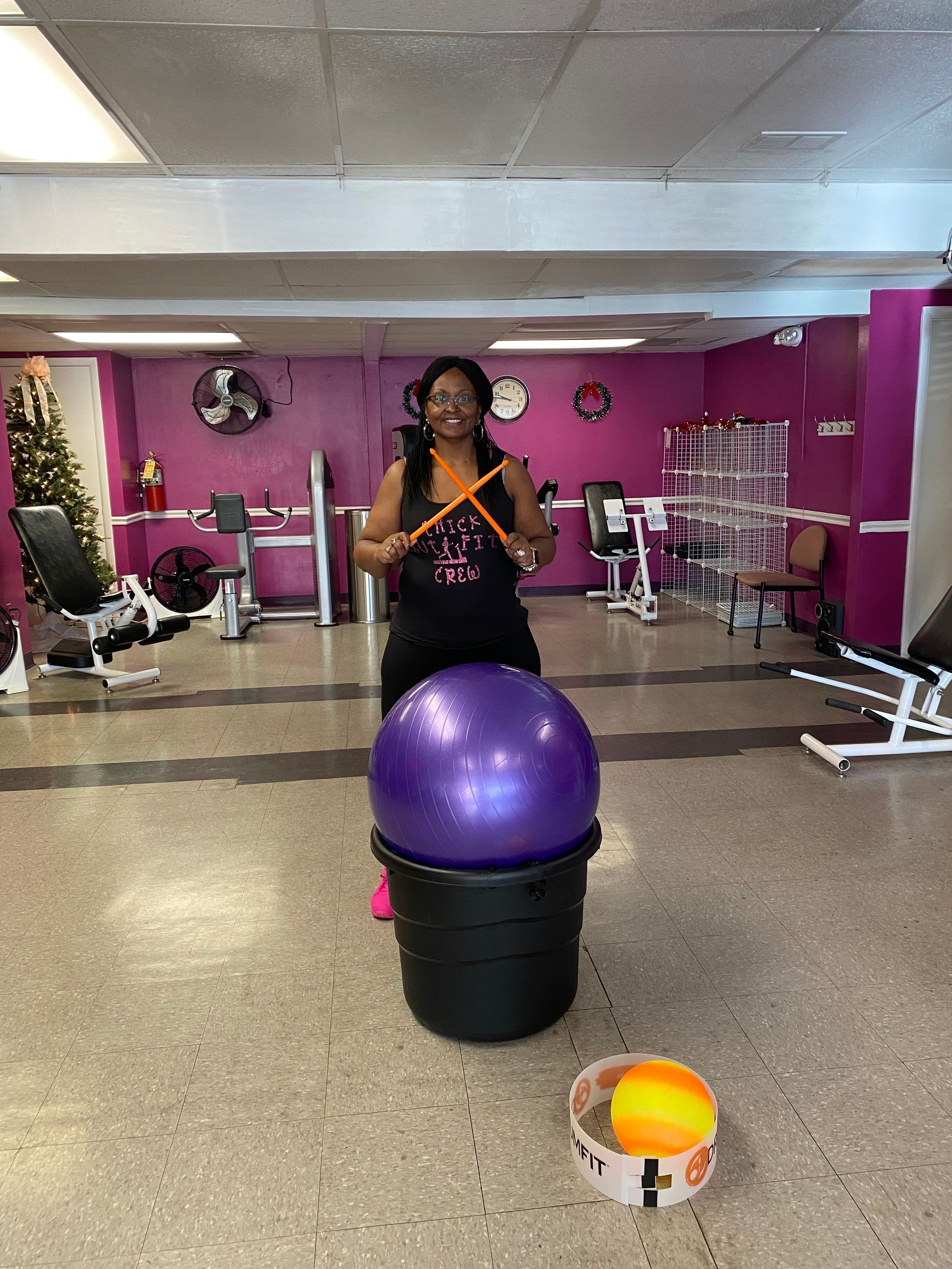 Woman in a purple gym standing behind a large exercise ball and holding a phone.
