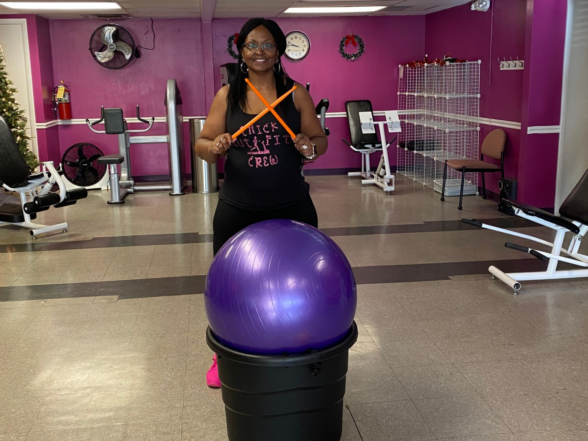 Person in a gym stands behind a purple exercise ball on a black stack, with fitness equipment in the background.