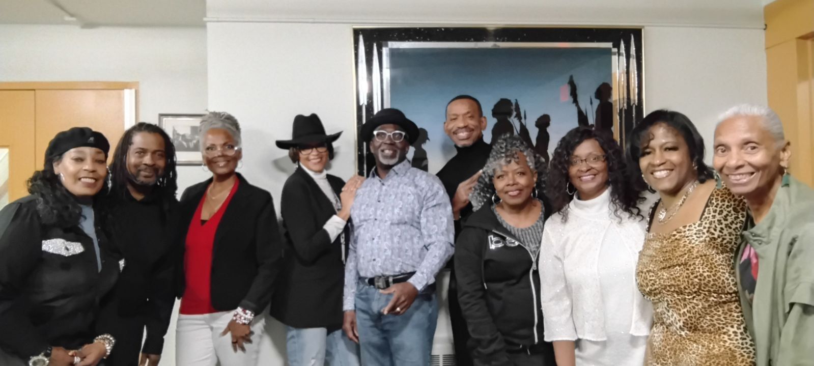Group of people posing indoors in front of a framed painting, smiling and standing close together.