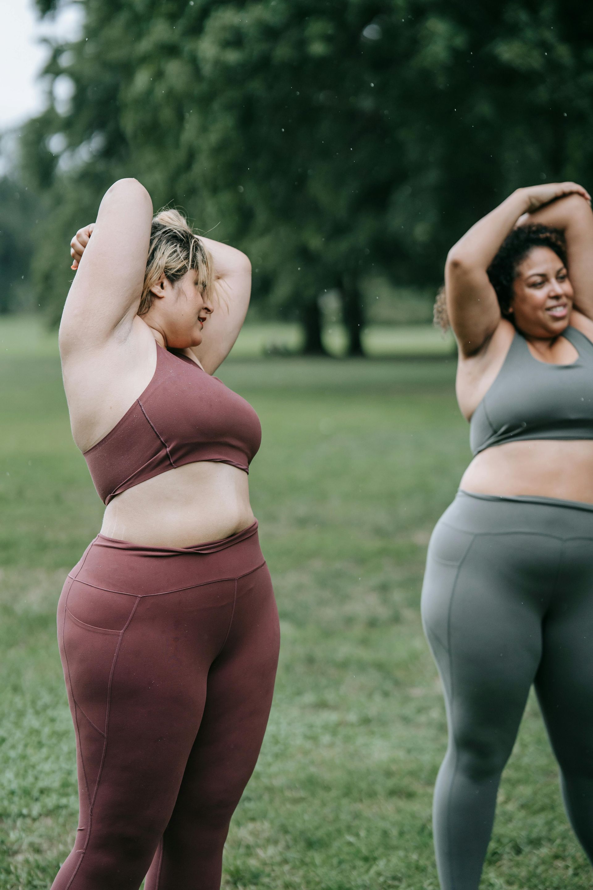 Two women stretching outdoors in workout clothes on a grassy field.