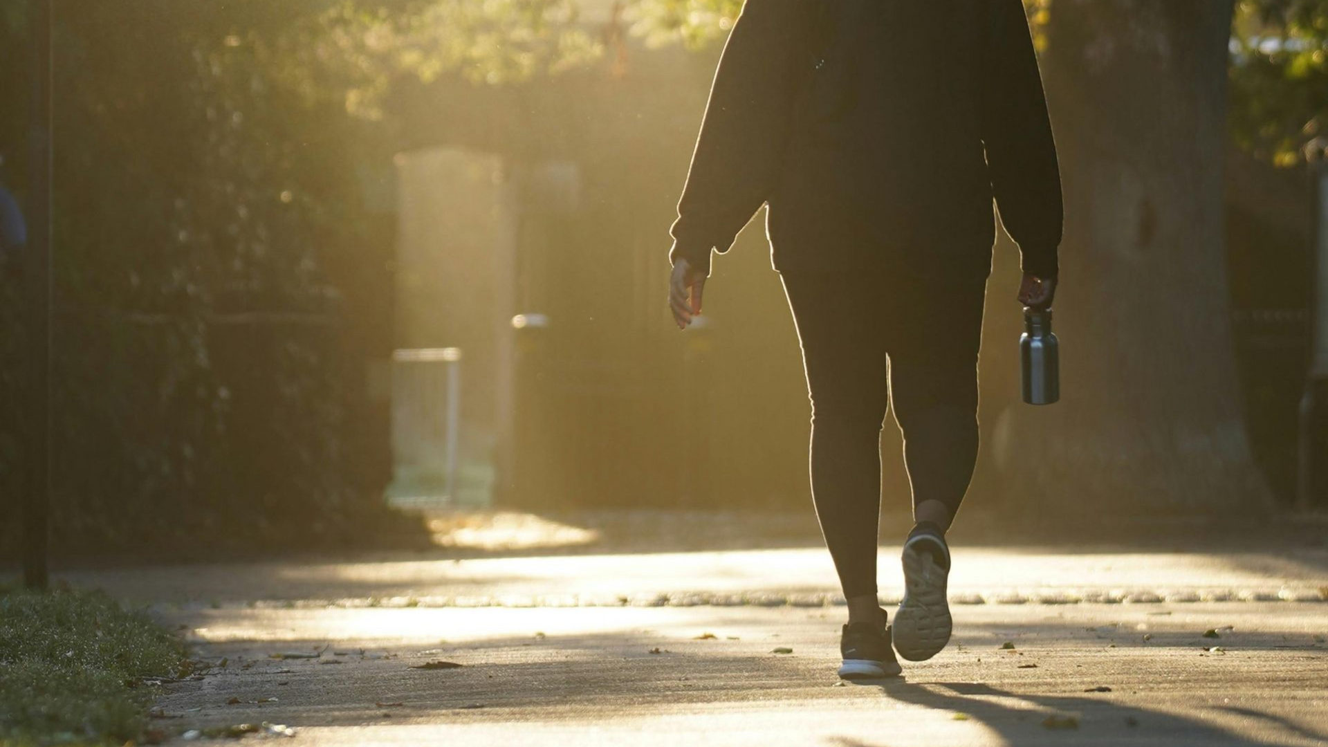 Person walking on a sunlit path carrying a water bottle, with long shadows in warm morning light