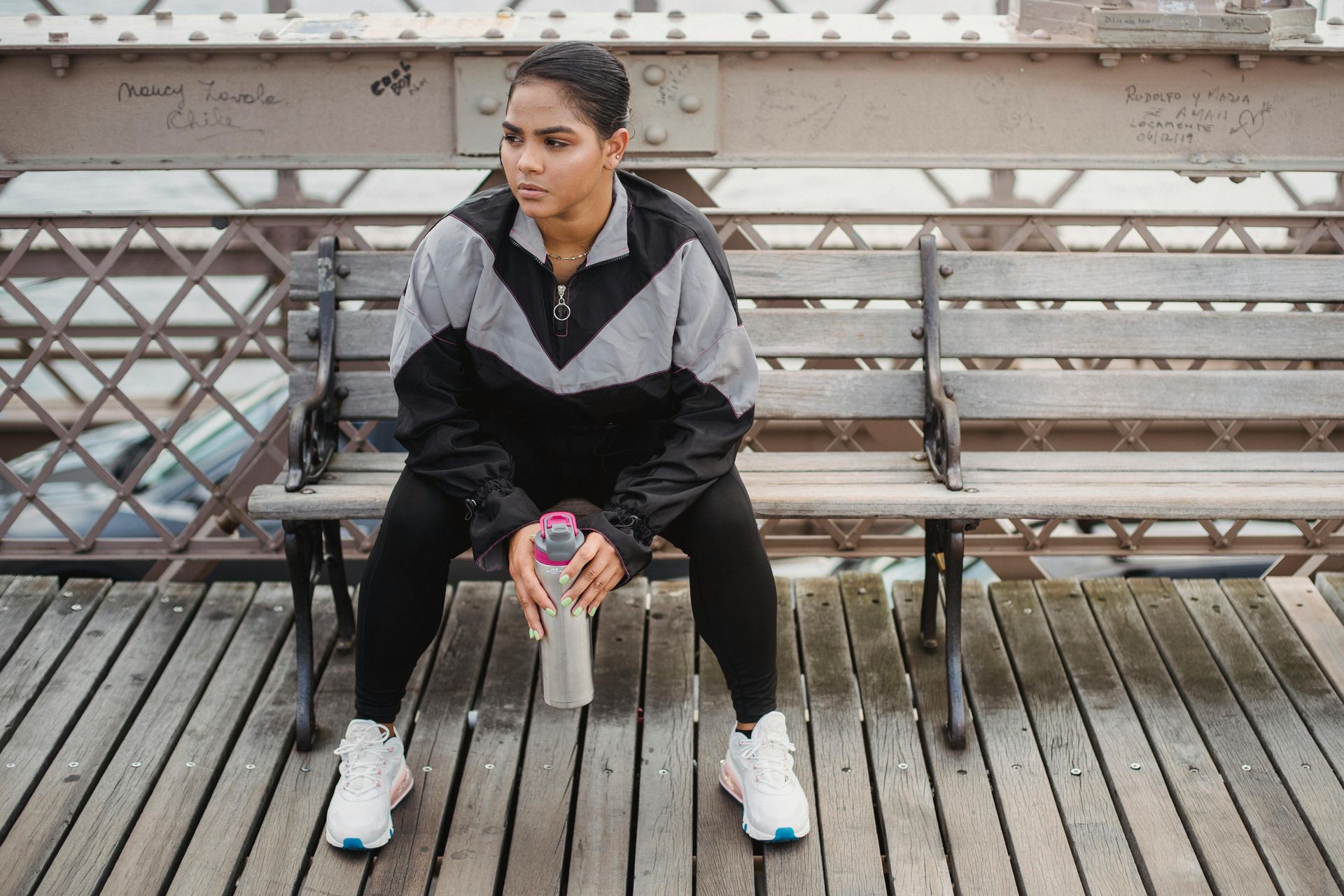 Person sitting on a bench holding a drink, wearing black and gray athletic clothes on a wooden deck.