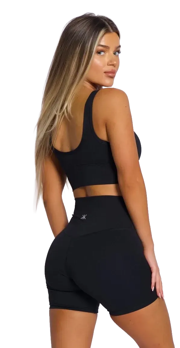 Woman in black athletic top and shorts posing against a white background, viewed from behind.