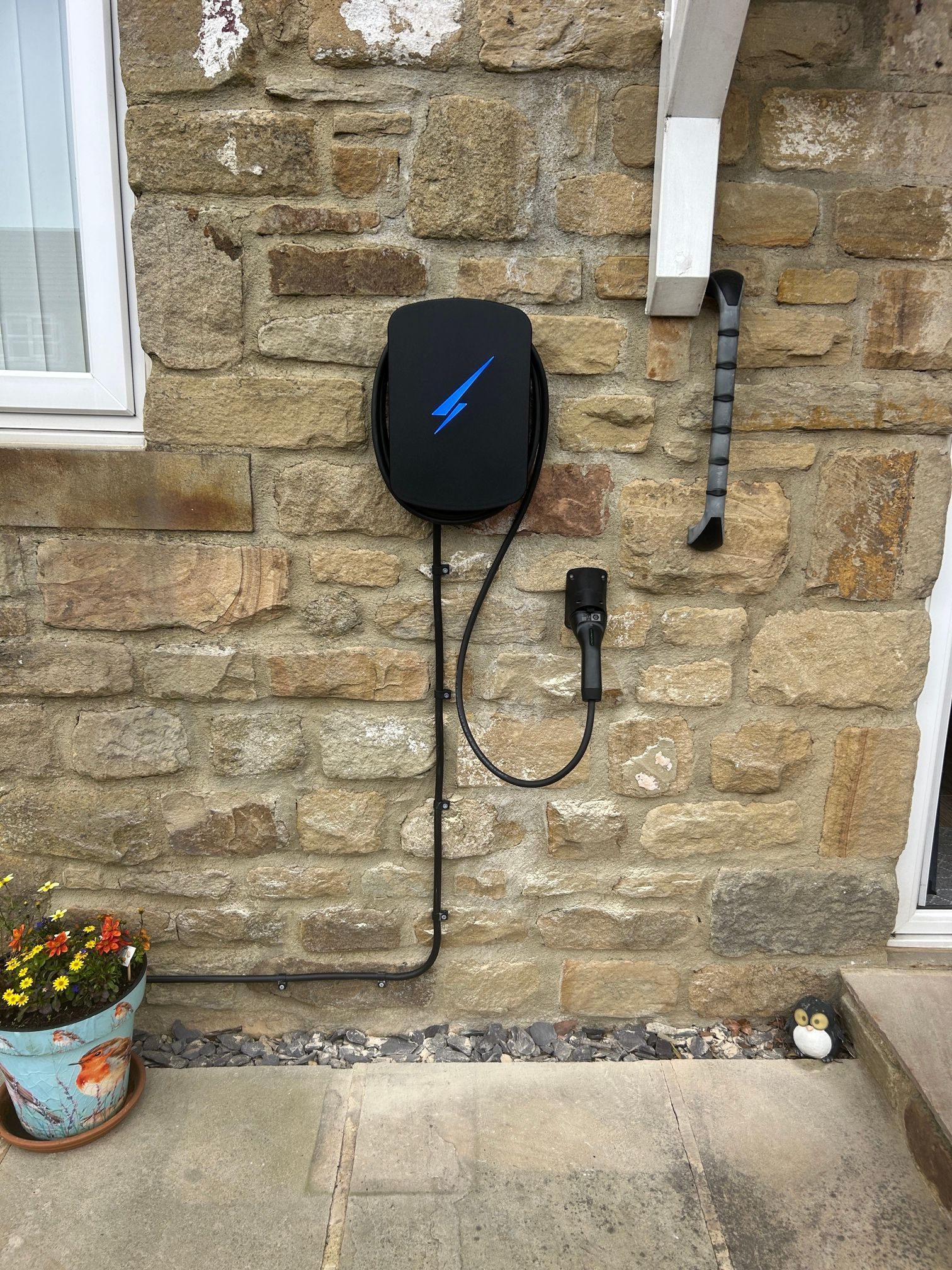 Black wall-mounted electric unit on a stone exterior wall, with cables and a small flower pot nearby