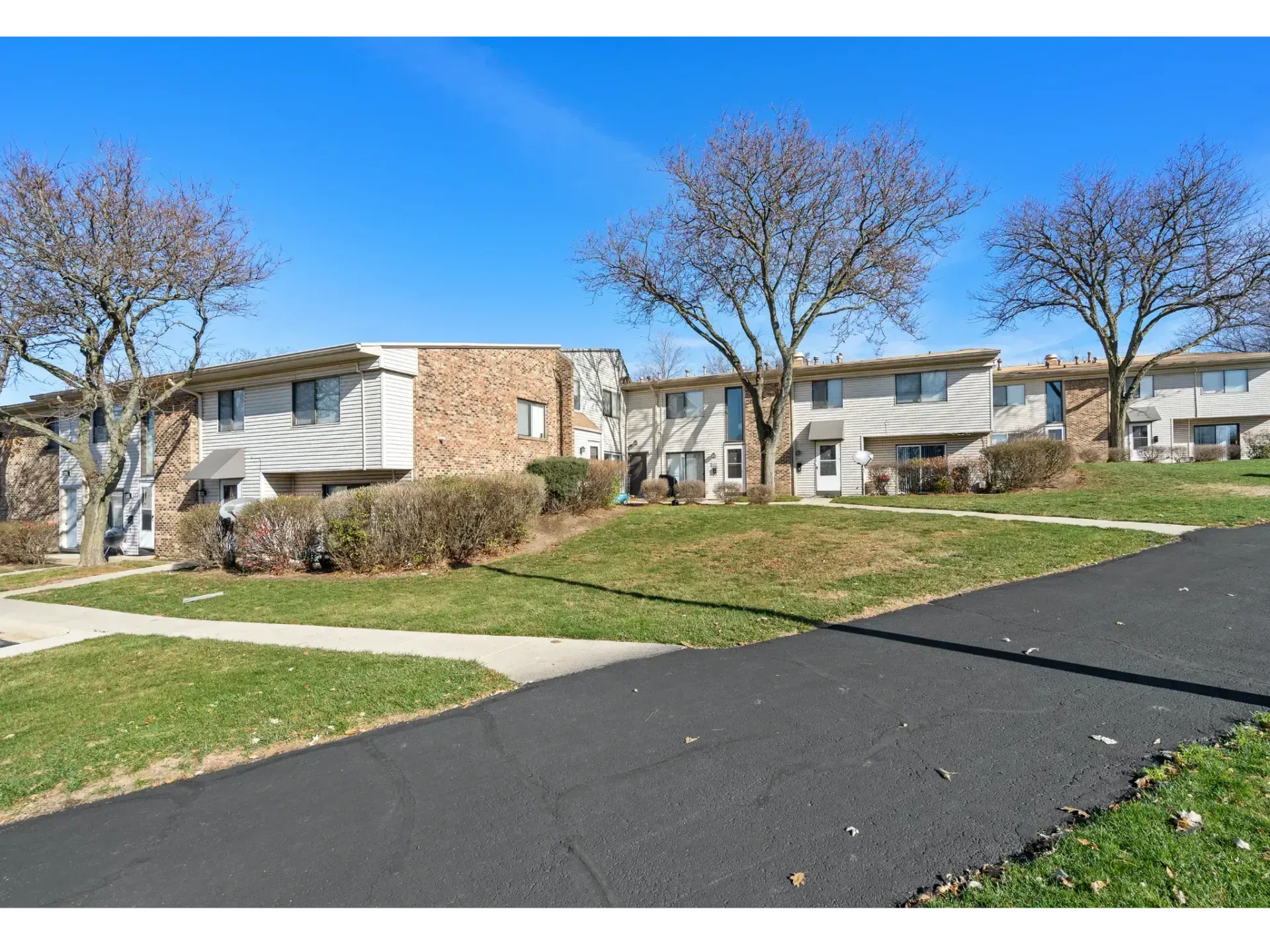 Apartment buildings beside a paved road with grass, leafless trees, and a clear blue sky